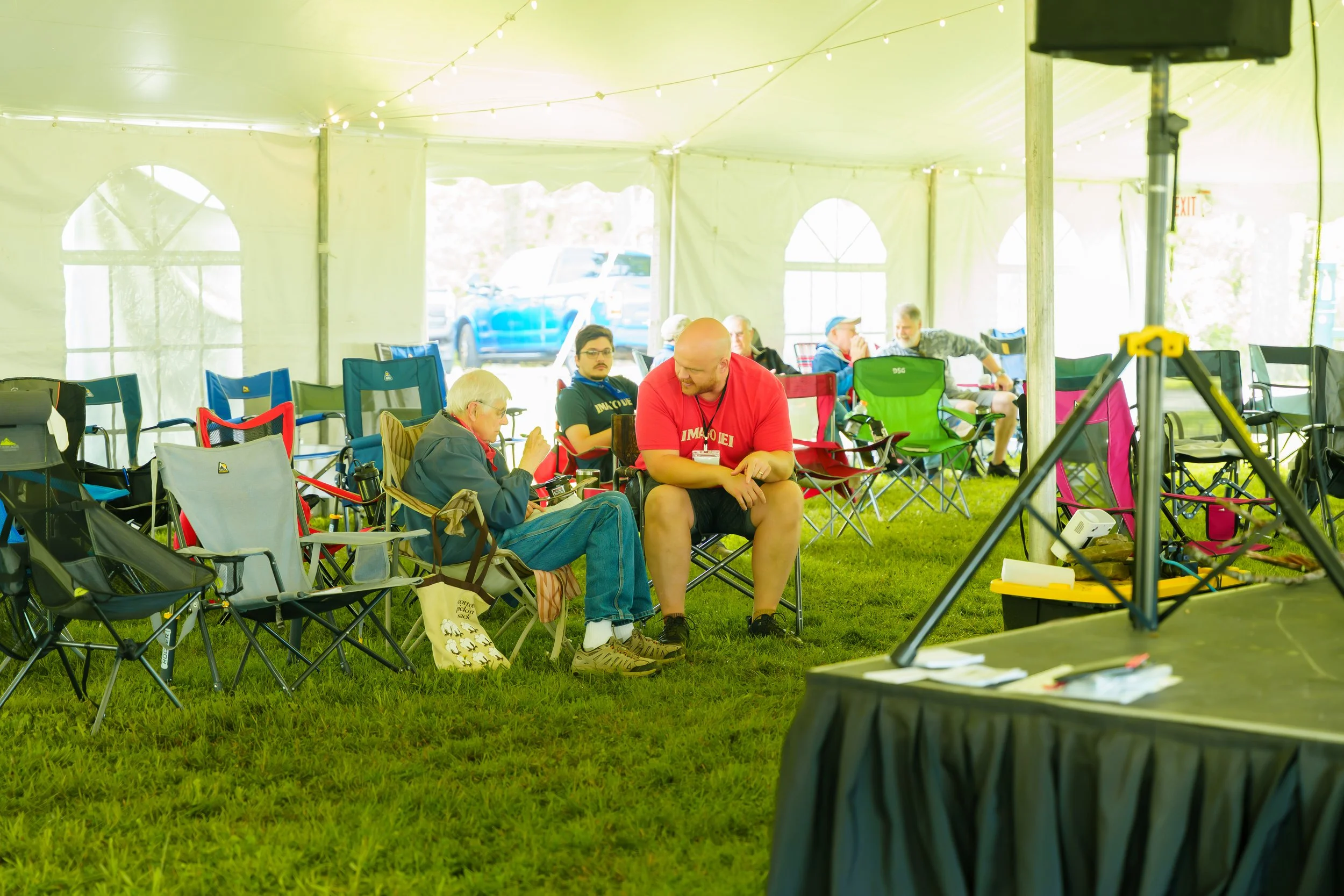 People sitting in camping chairs inside a large white event tent with string lights, grass flooring, and several windows, during daytime.