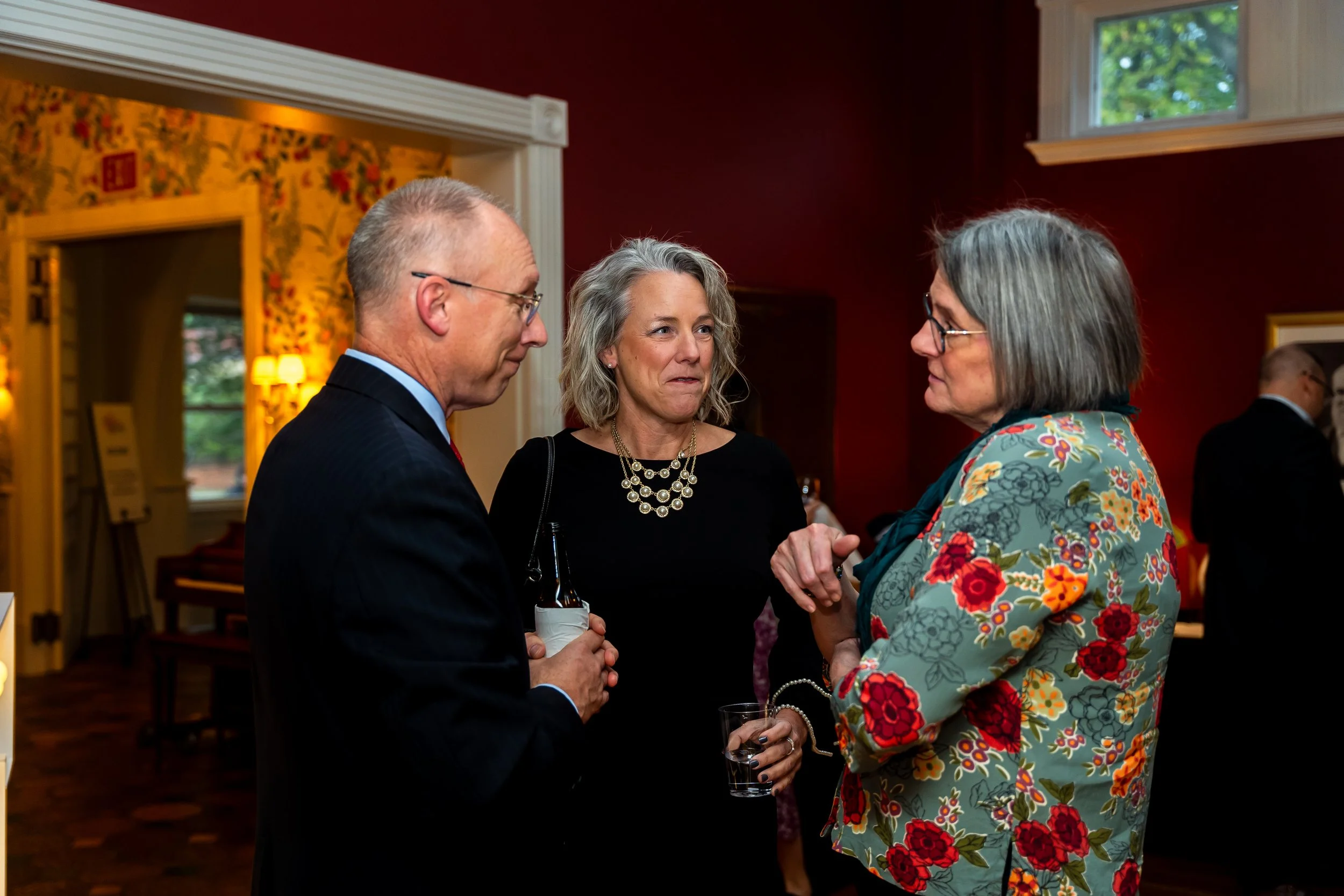 Three people in formal attire are engaged in conversation at a social event in a warmly lit room with red walls and floral curtains. An older woman with gray hair, wearing a floral blazer and glasses, is holding a glass of drink. A man with glasses a