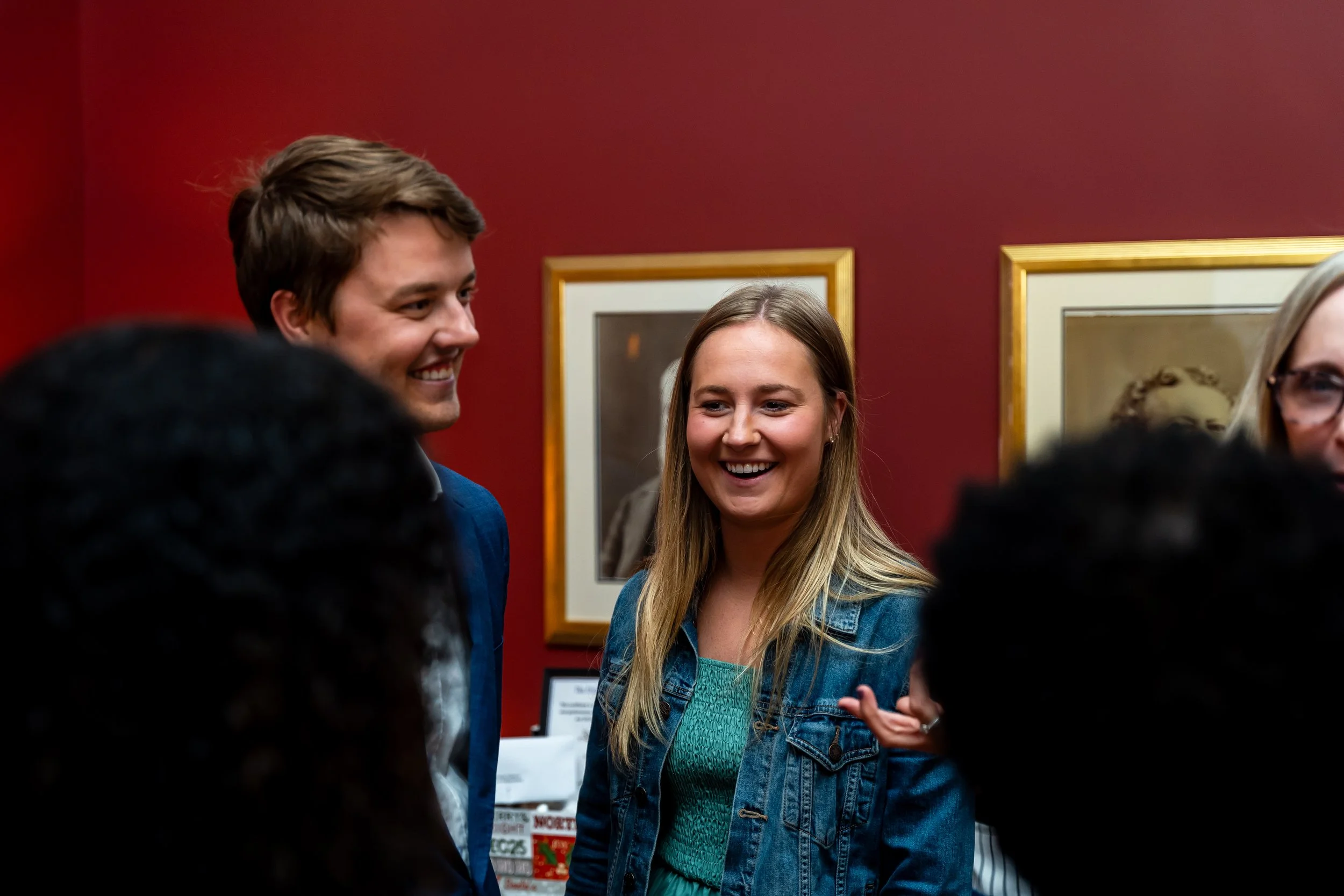 People at an art gallery or museum engaging in conversation. Two individuals, a man and a woman, are smiling and talking to each other, surrounded by framed artwork on a red wall.