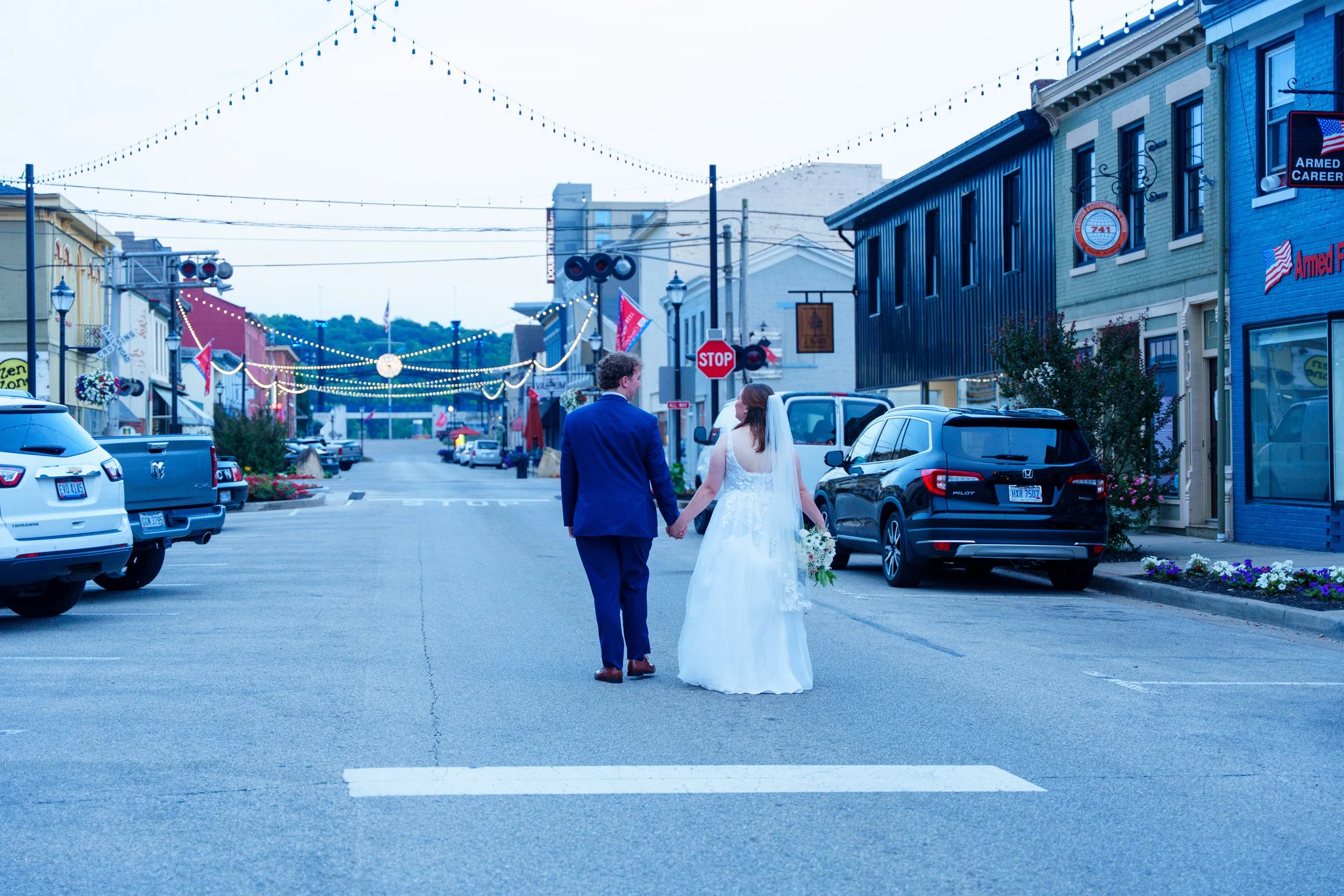 A bride and groom holding hands walking across a city street decorated with string lights during early evening.