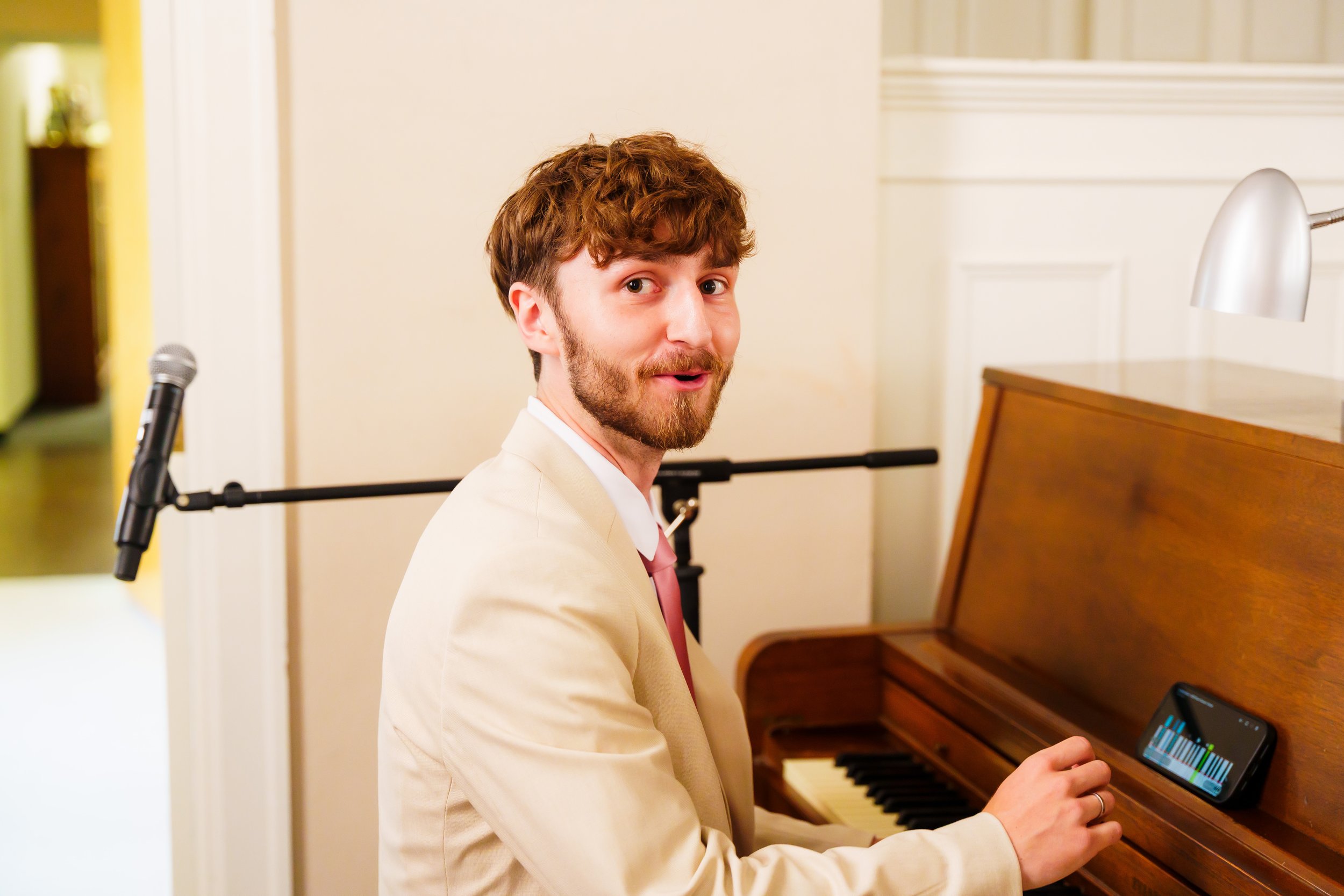 A man with curly red hair and a beard wearing a white blazer and pink tie, sitting at a wooden piano, looking over his shoulder at the camera, holding a smartphone with a music app displayed on the screen.