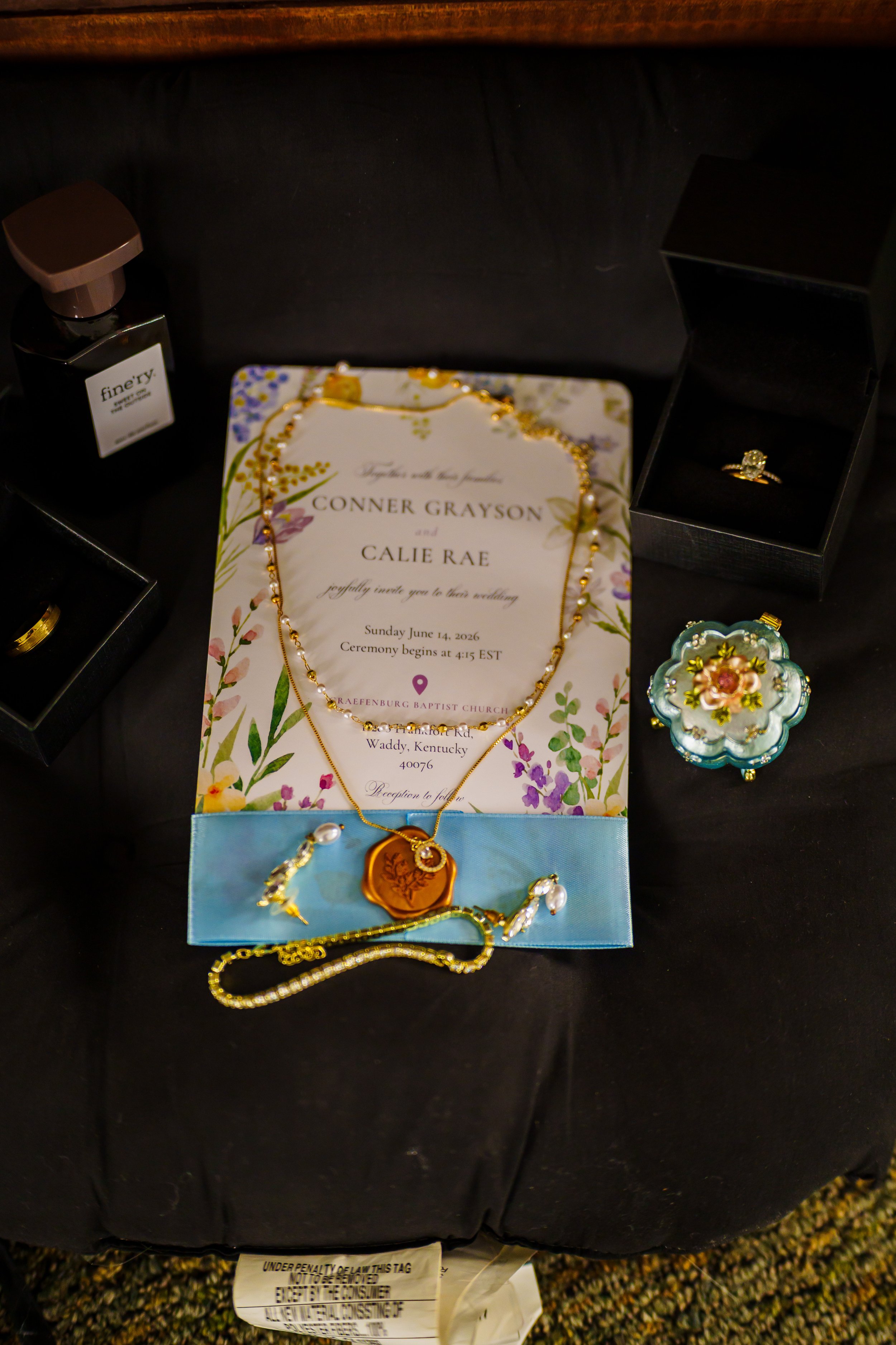 A black surface with an open jewelry box containing a ring with a large central stone, a small jewelry box with decorative floral design, a greeting card for a wedding with floral design and text, and other jewelry pieces including necklaces and brac