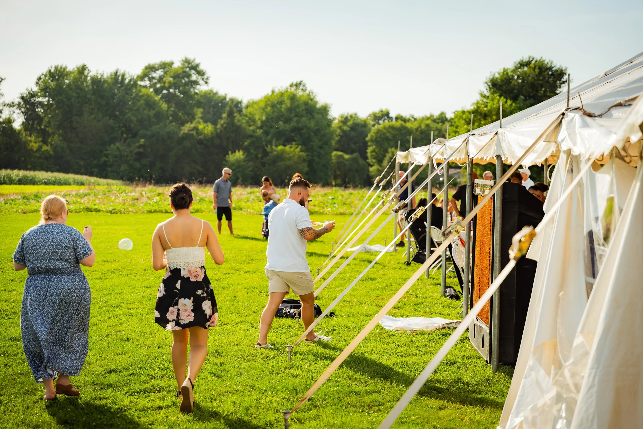 People at an outdoor event with a white canopy tent, green grassy field, trees, and clear sky.