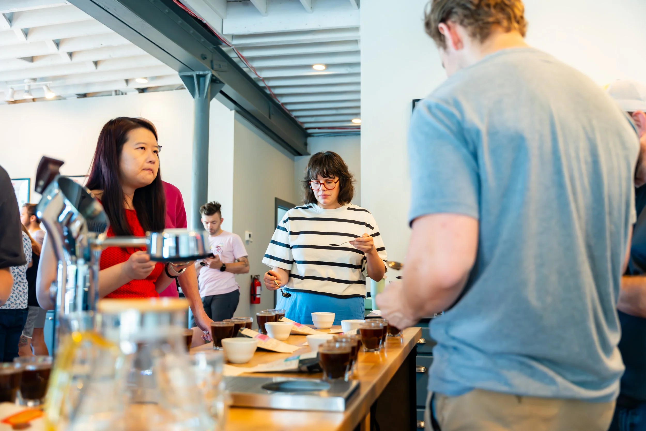 People participating in a coffee tasting event, with various coffee cups on a table and a barista preparing coffee in a modern indoor setting.