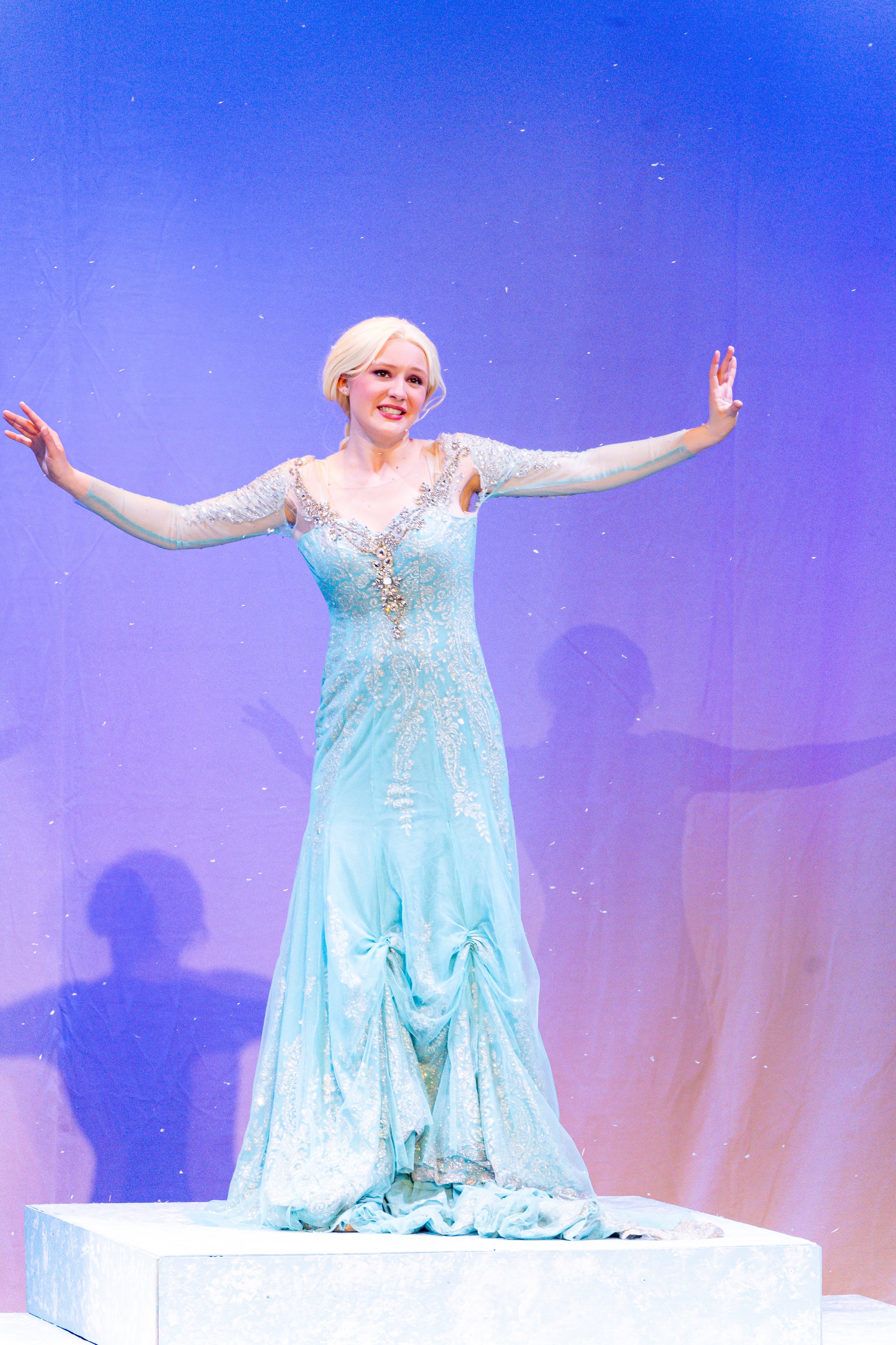A woman dressed as Elsa from Disney's Frozen, wearing a light blue gown with silver embellishments, standing on a white platform with her arms outstretched against a purple backdrop.
