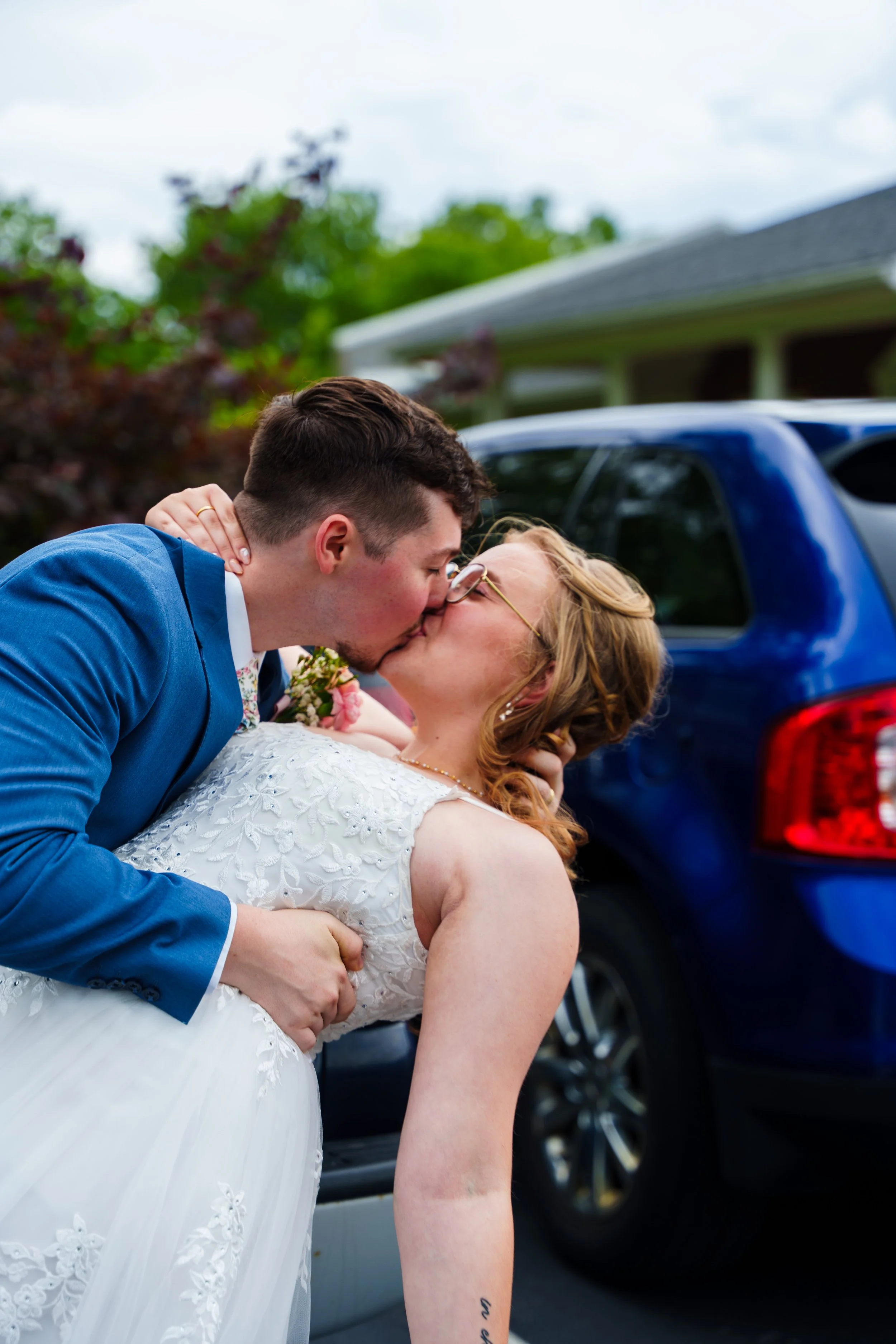 A groom in a blue suit kisses a bride in a white wedding dress, holding her in a dip outside near a blue vehicle.