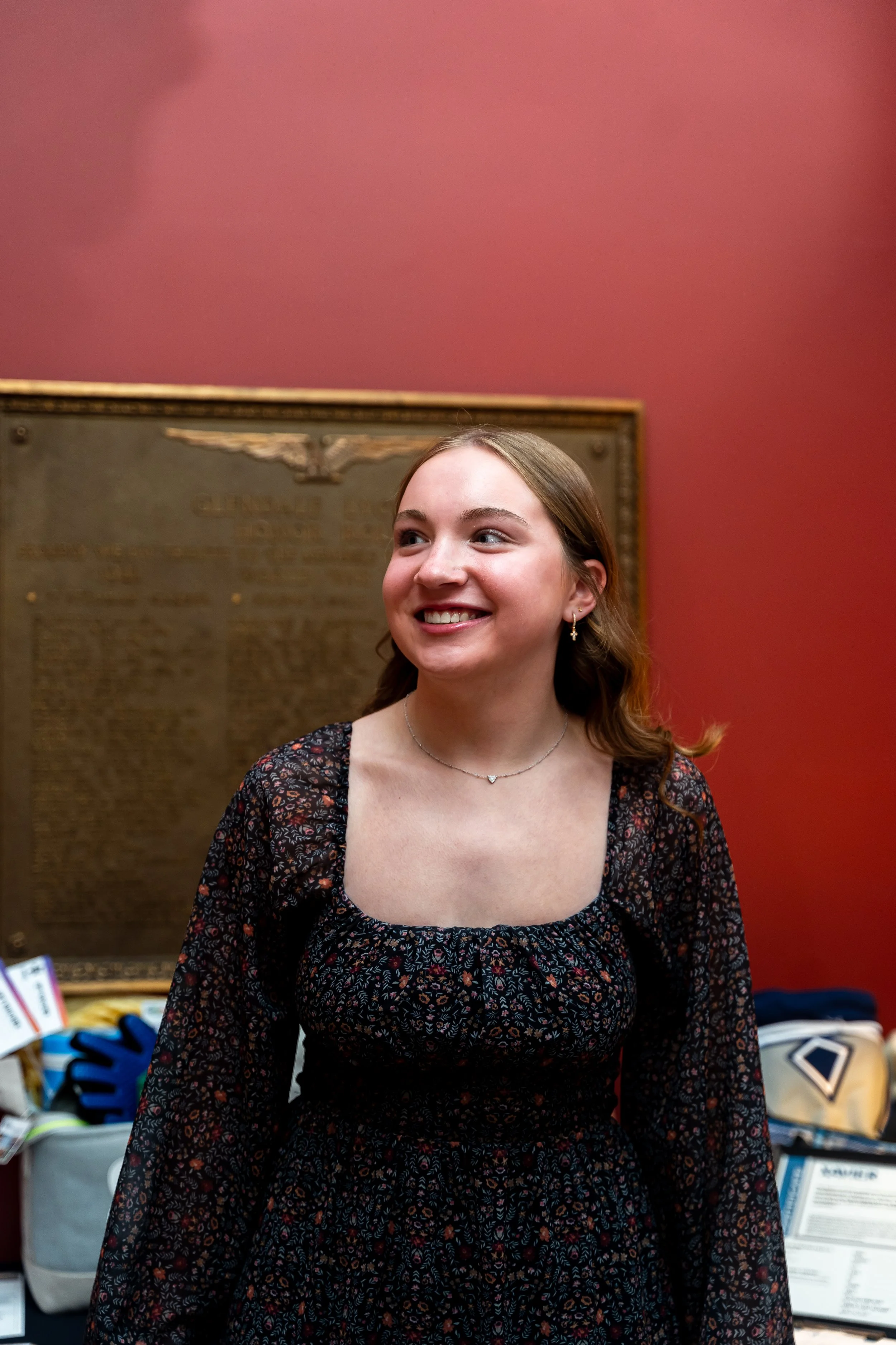 Young woman with brown hair smiling, wearing a black floral dress and a necklace, standing in front of a framed plaque on a red wall.