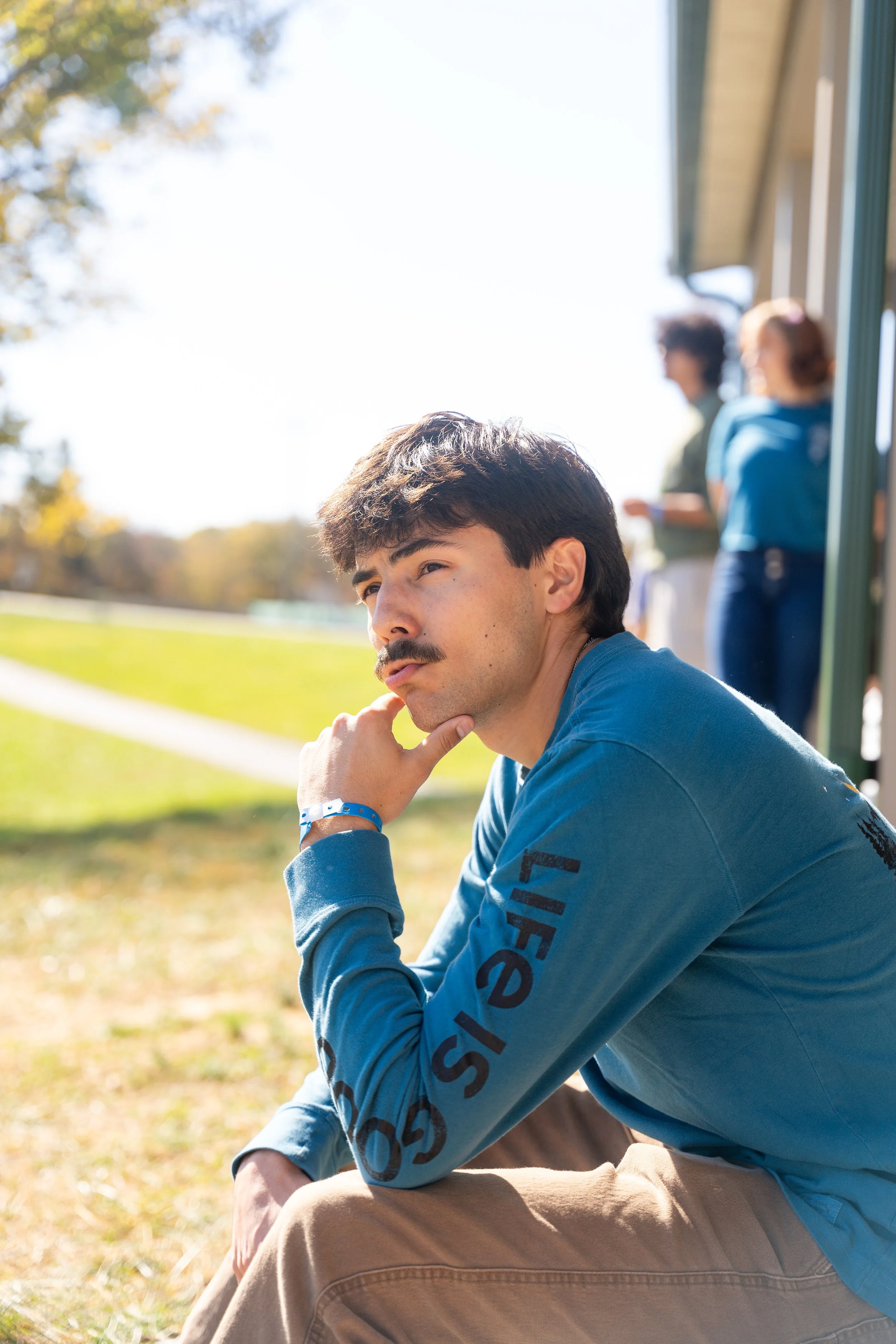 A young man with dark hair and a mustache sitting outdoors on grass, resting his chin on his hand, wearing a blue long-sleeve shirt and khaki pants, with other people blurred in the background.