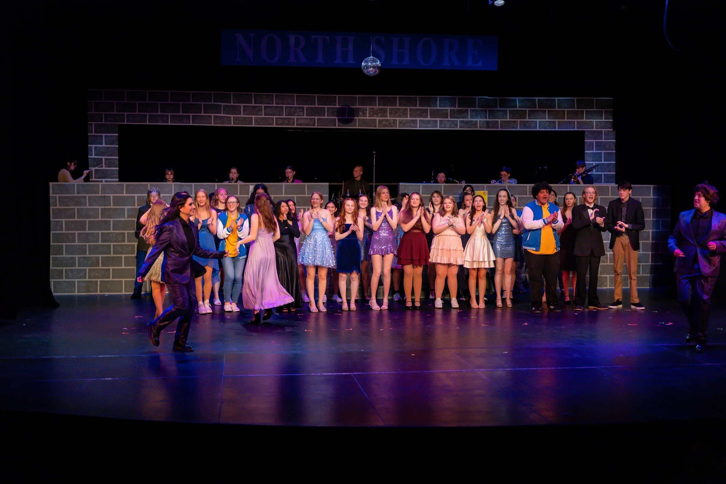 A group of young performers on stage dressed in colorful costumes, some dancing, with a backdrop that reads 'North Shore' and a small disco ball hanging above.