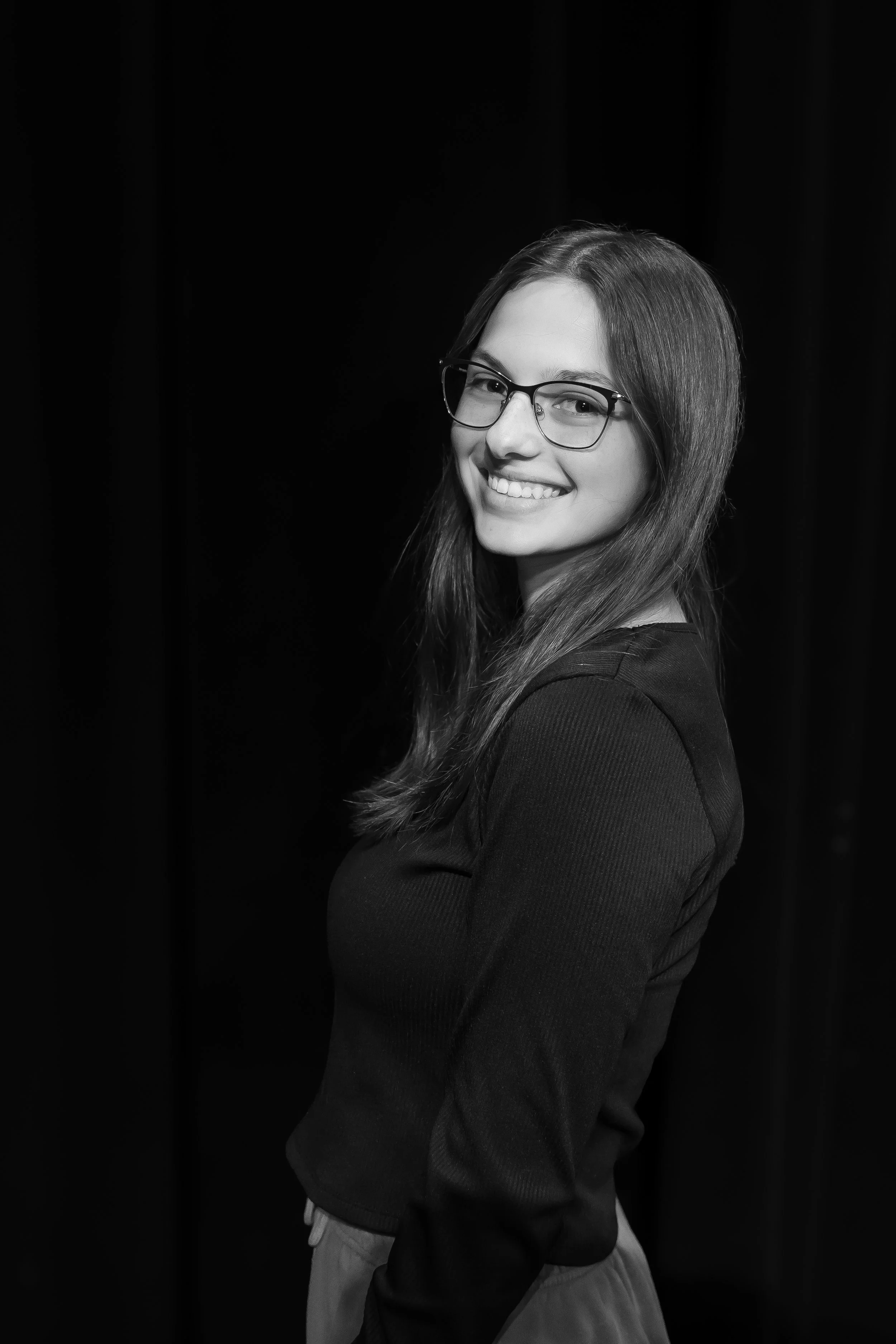 A young woman with glasses and long hair, smiling and wearing a dark top, standing against a plain dark background.