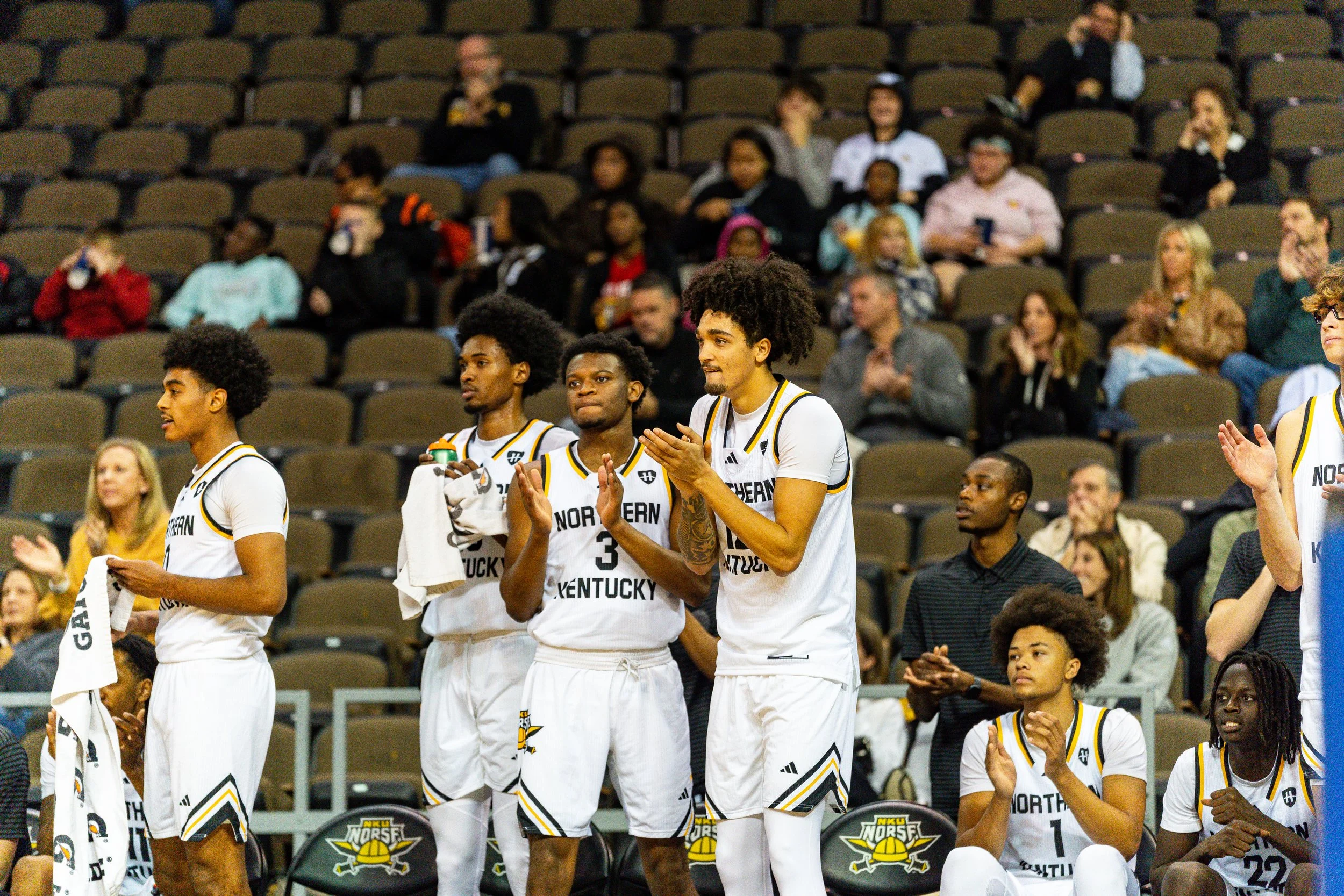 Basketball players on the court during a game, wearing white jerseys with 'Northern Kentucky' written on them, some clapping and others sitting on the bench, with spectators in the background.