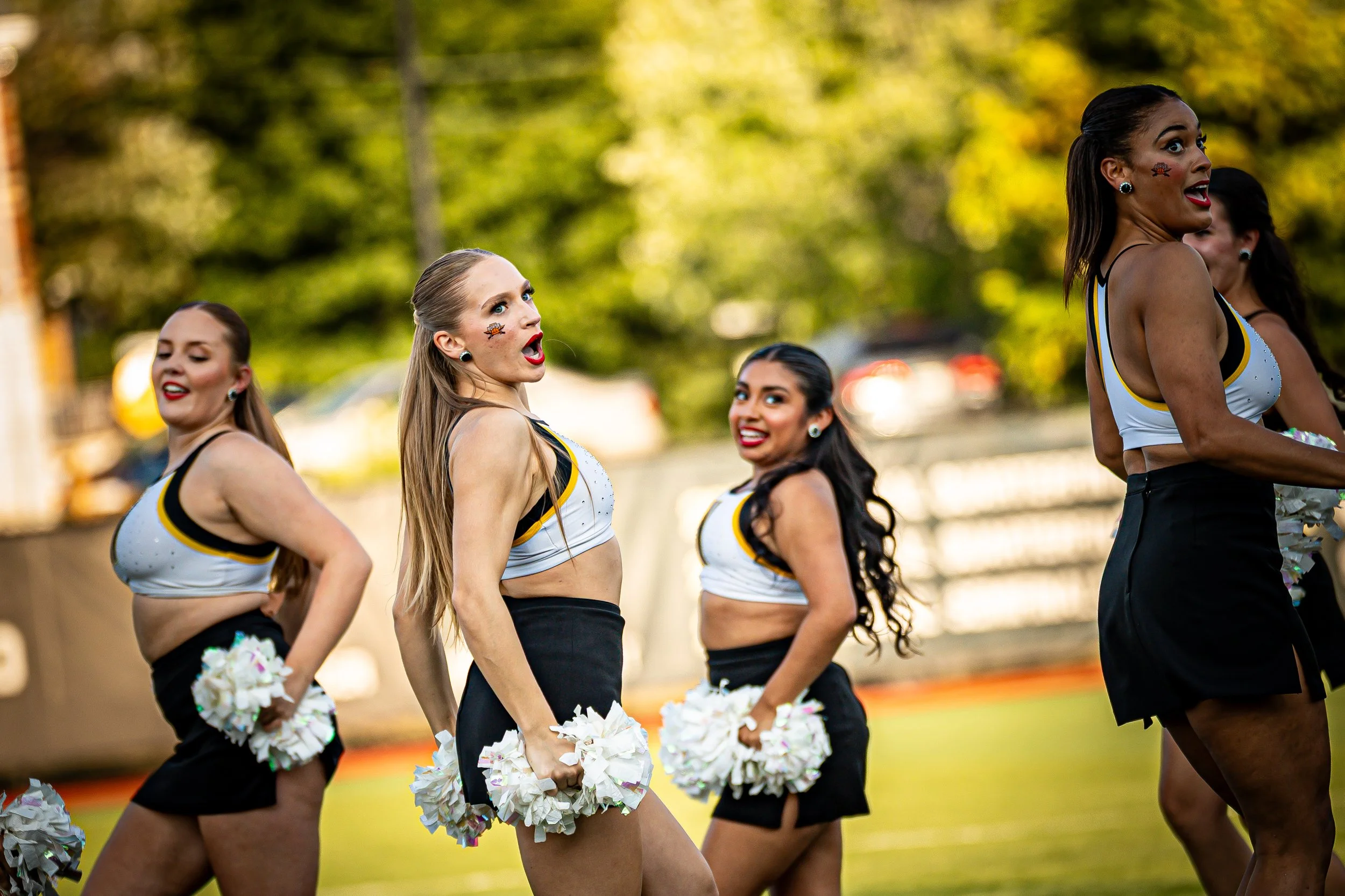 A group of cheerleaders performing a dance routine outdoors on a football field during daytime, wearing matching uniforms with white tops and black skirts, holding white pom-poms.