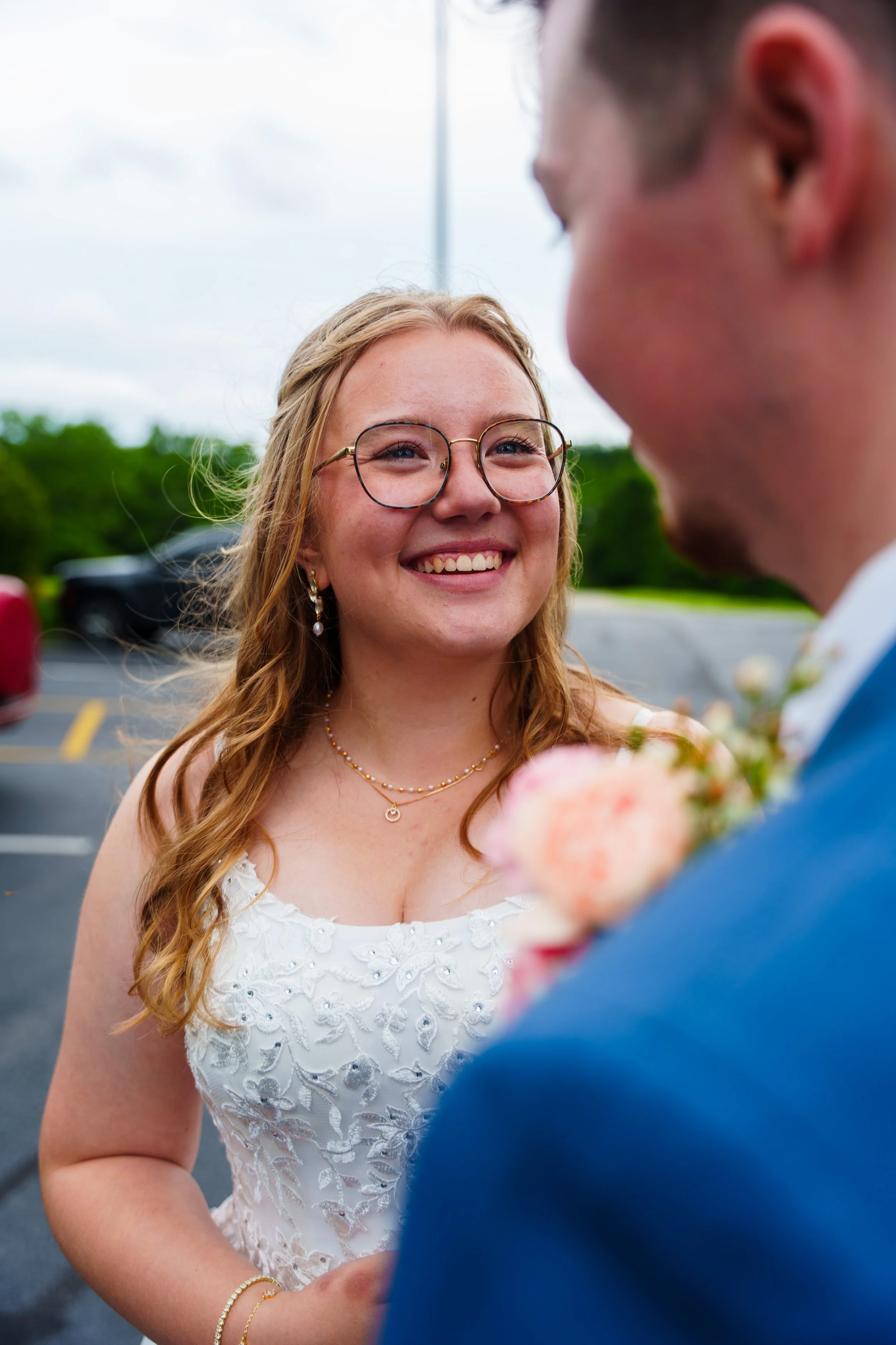 A woman with red hair and glasses smiling at a man in a blue suit during a outdoor wedding ceremony, with green trees and parked cars in the background.
