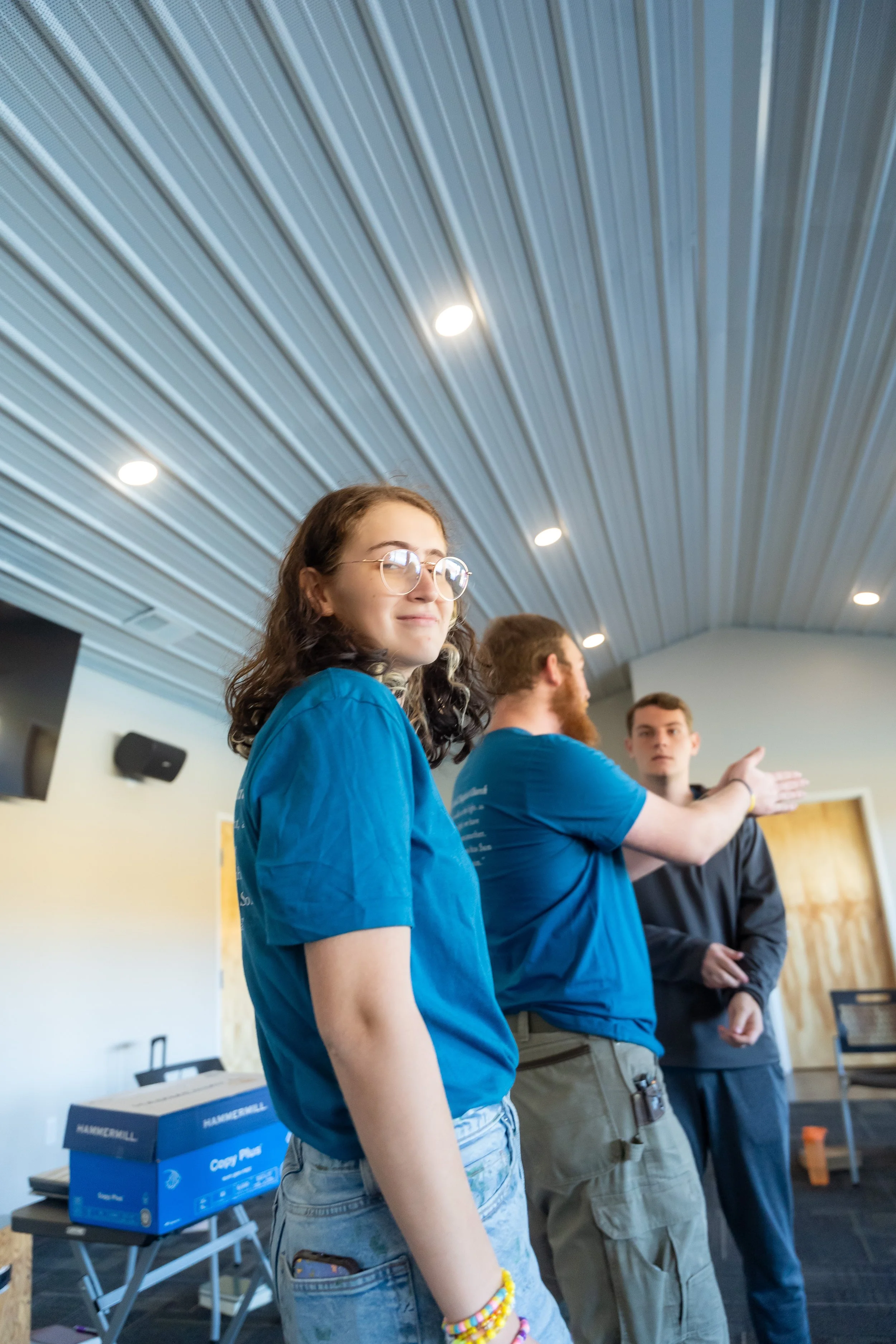 A group of young people standing and talking in a room with a corrugated metal ceiling and recessed lighting. The foreground features a girl with glasses wearing a blue shirt and colorful bracelets, with other individuals in the background.