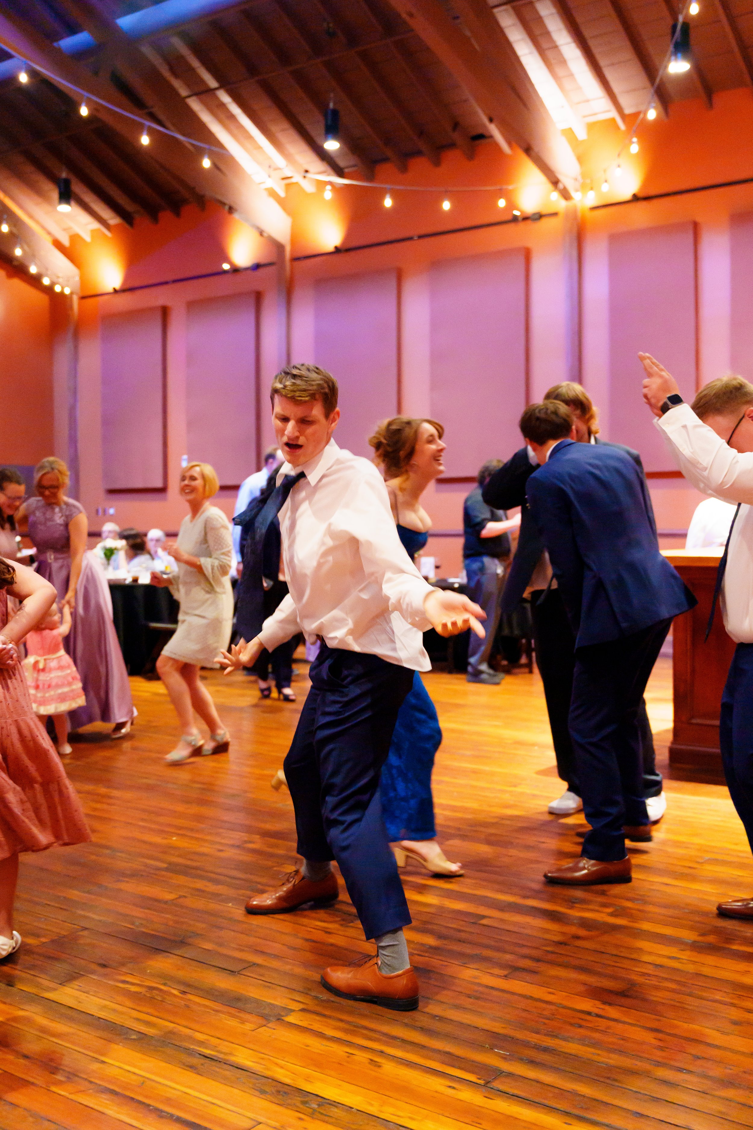 People dancing at a party in a wooden-floored hall with string lights and pink walls.