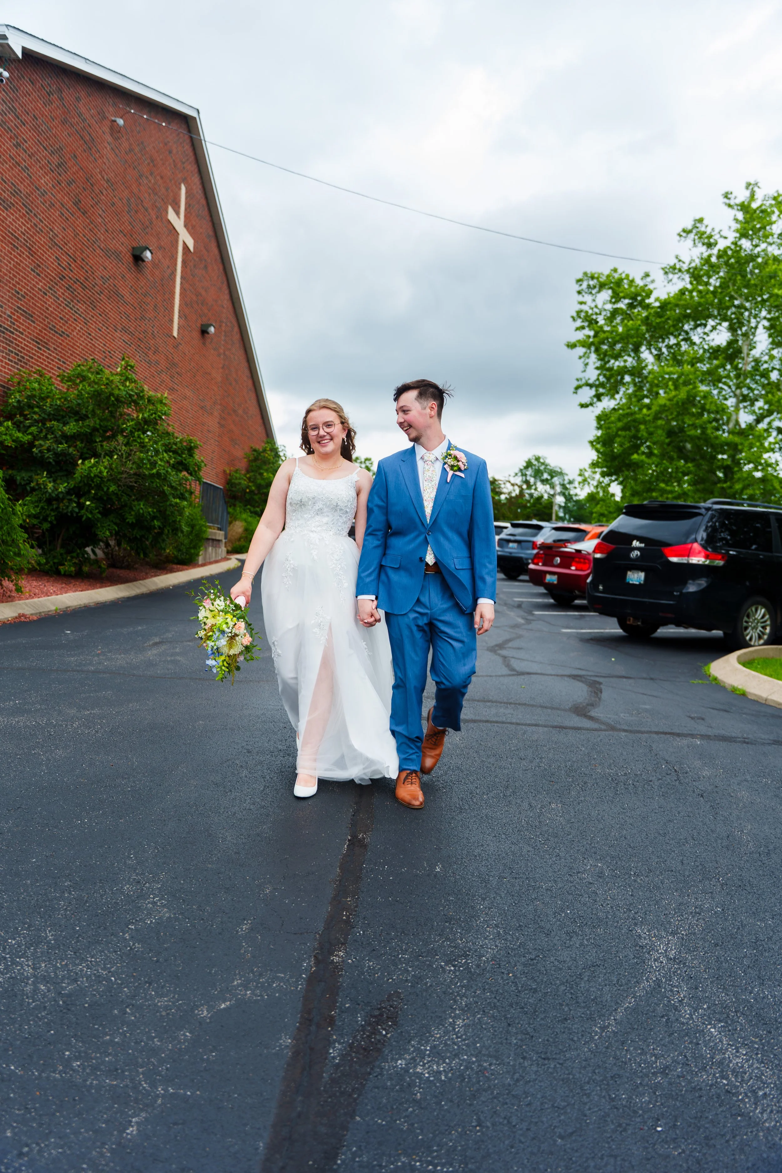 A bride and groom walking hand in hand outside a church with a large white cross on the brick wall, smiling and holding a bouquet of flowers.