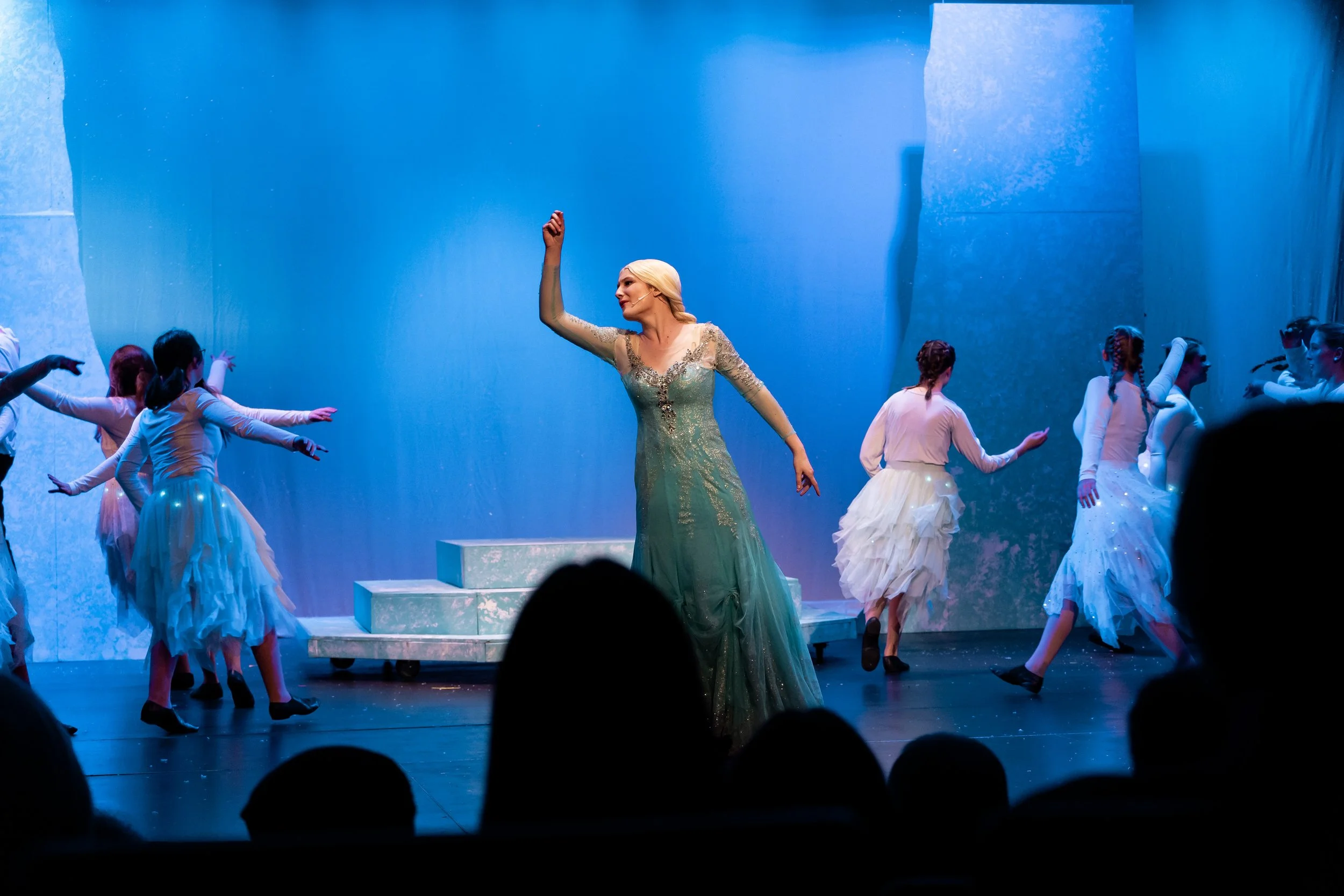 A stage scene from a theatrical performance featuring a woman in a green, glittery gown with long blonde hair, surrounded by dancers in white costumes with ruffled skirts, all performing a dance routine against a blue background.