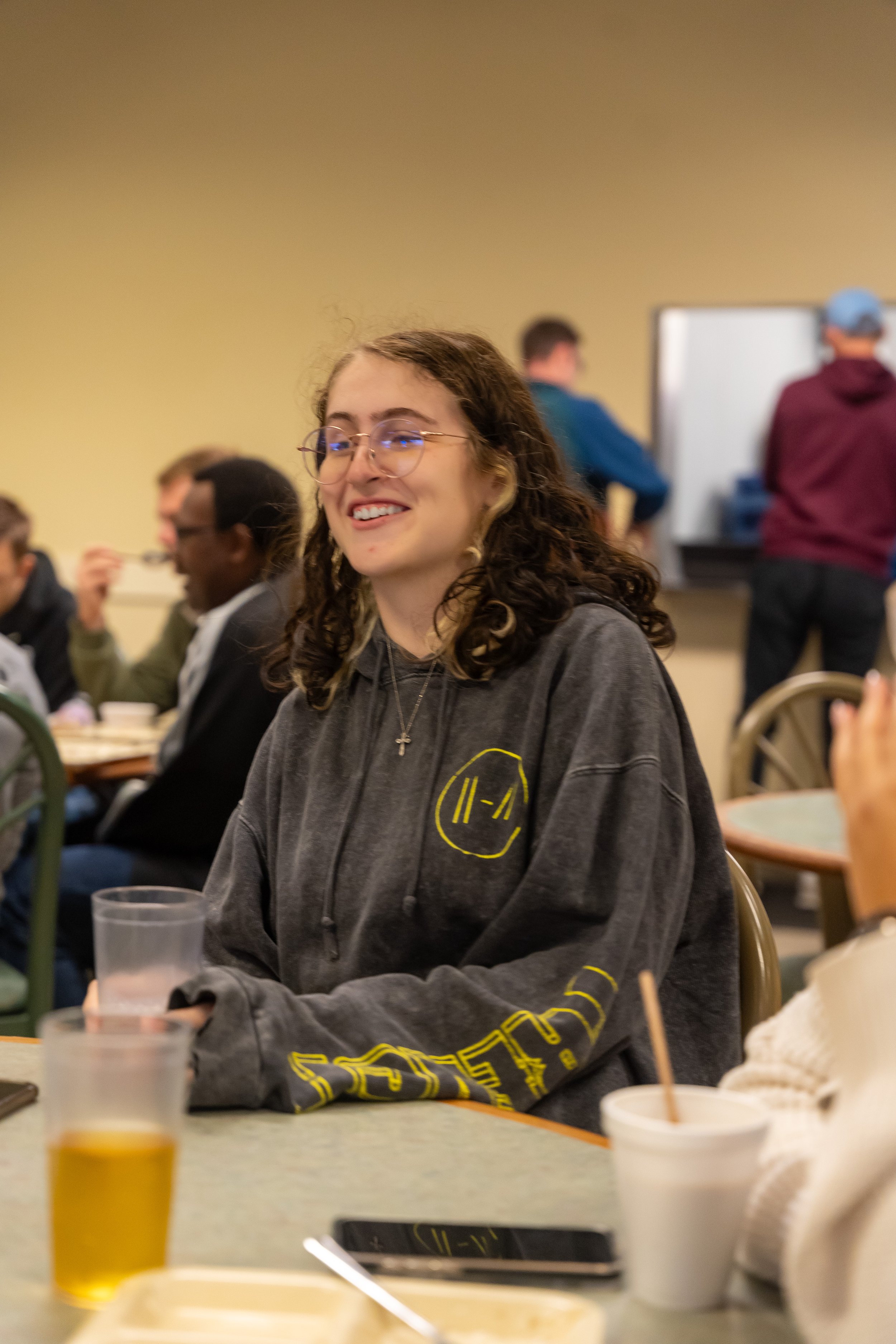 Young woman with curly hair and glasses smiling at a social gathering, seated at a table with drinks and food, in a room with beige walls and other people in the background.