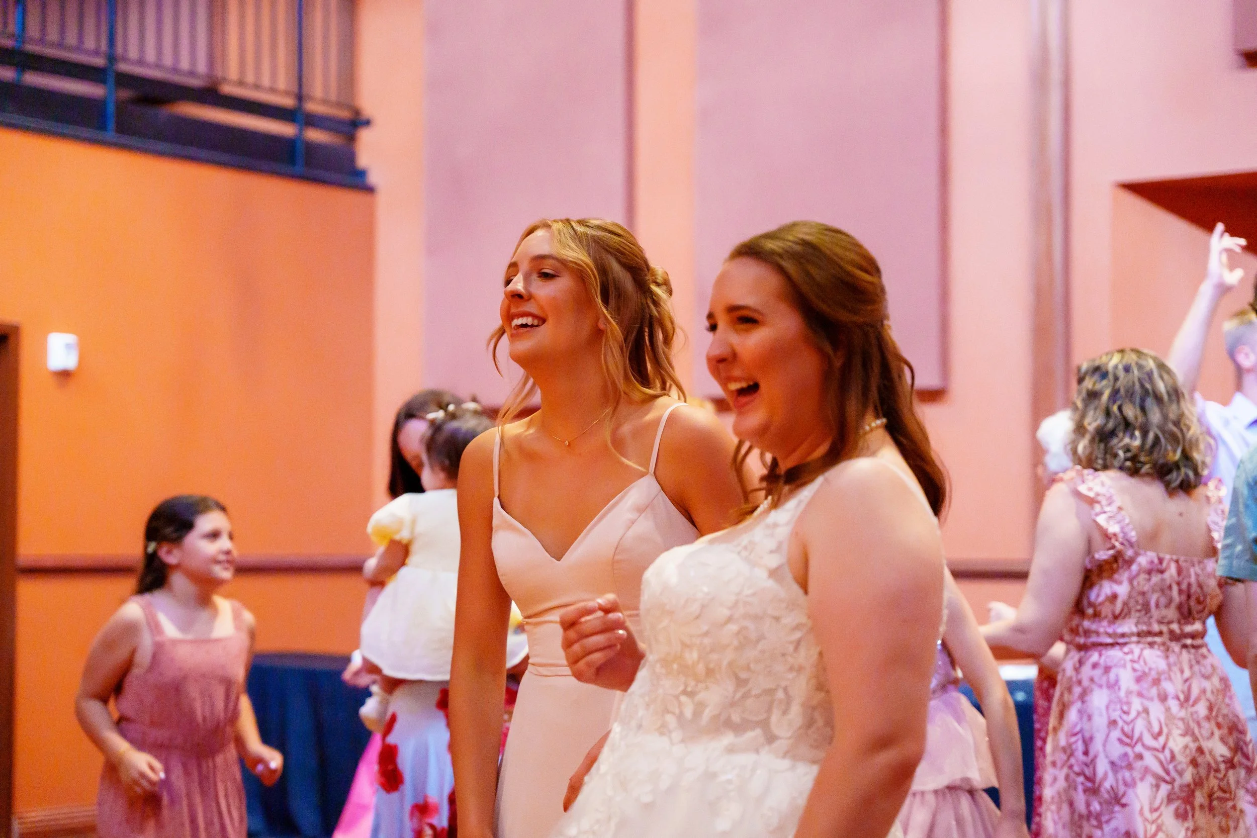 Two young women in white dresses dancing at a party, surrounded by other children in colorful dresses in a decorated indoor hall.