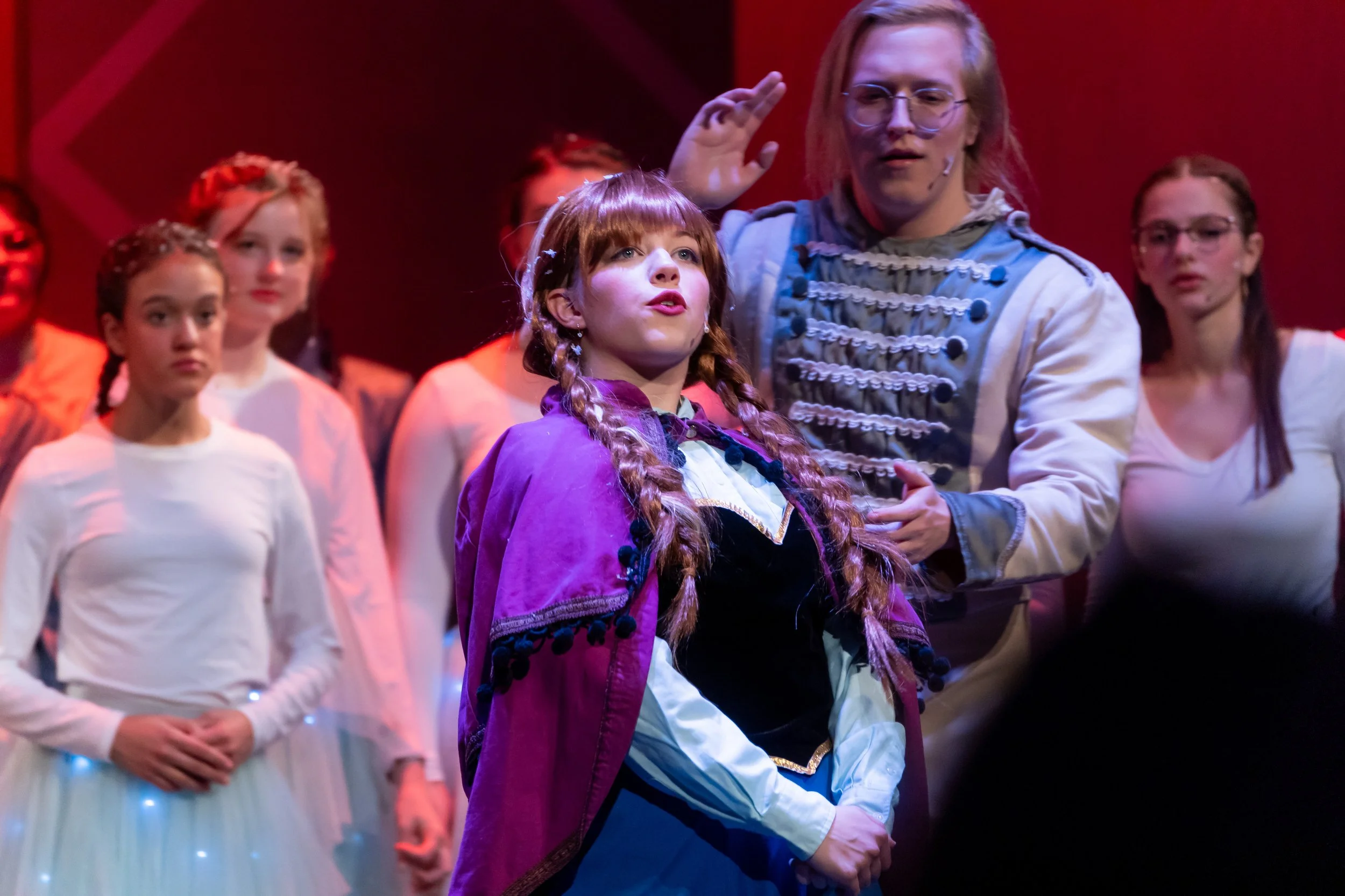 Scene from a stage production with children and an adult in costume, character likely from a fairytale, with a passionate expression, audience or cast members in background, red-lit background.