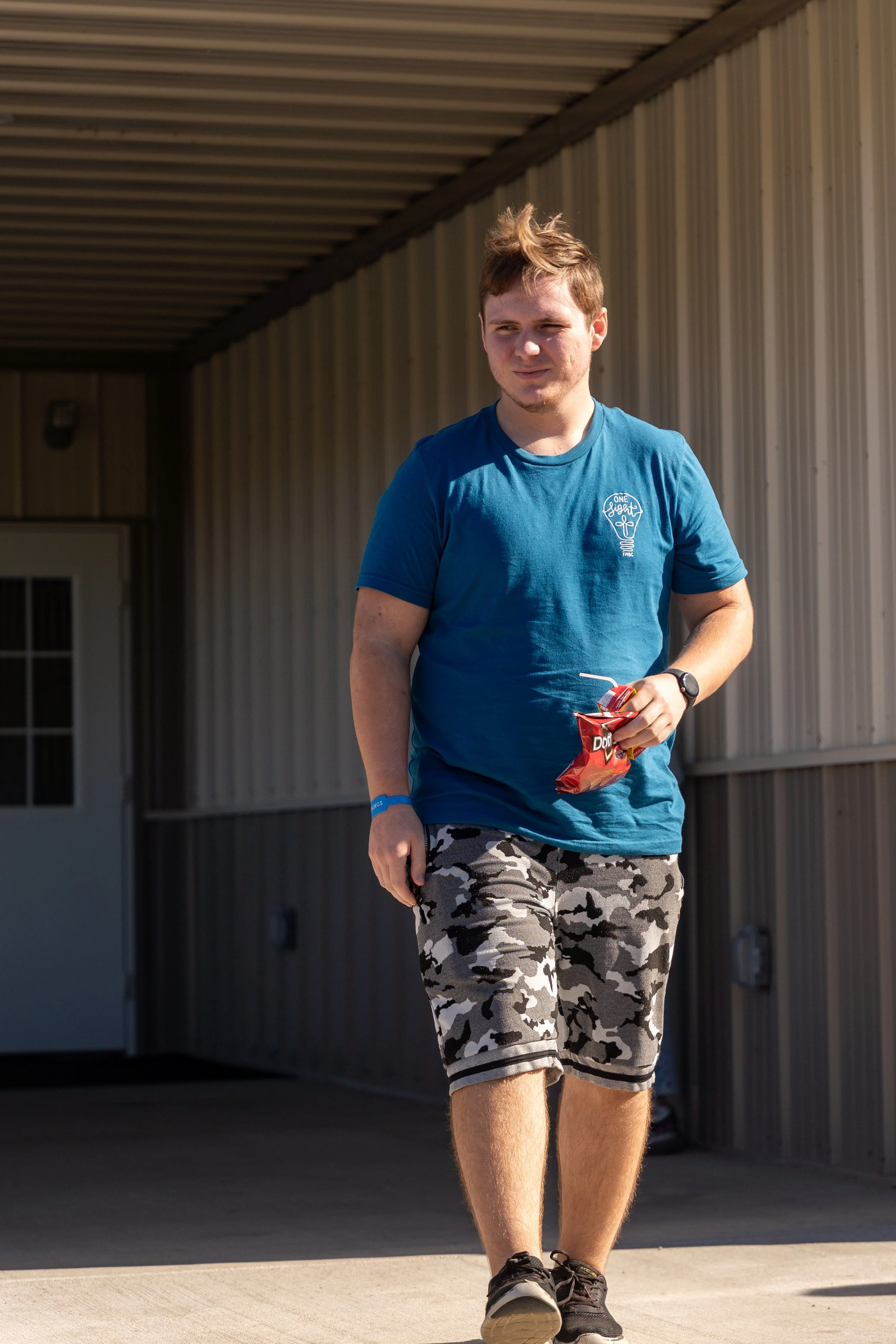 A young man with light brown hair walking outdoors, wearing a blue T-shirt, camouflage shorts, and black sneakers, holding a can of soda and a snack.
