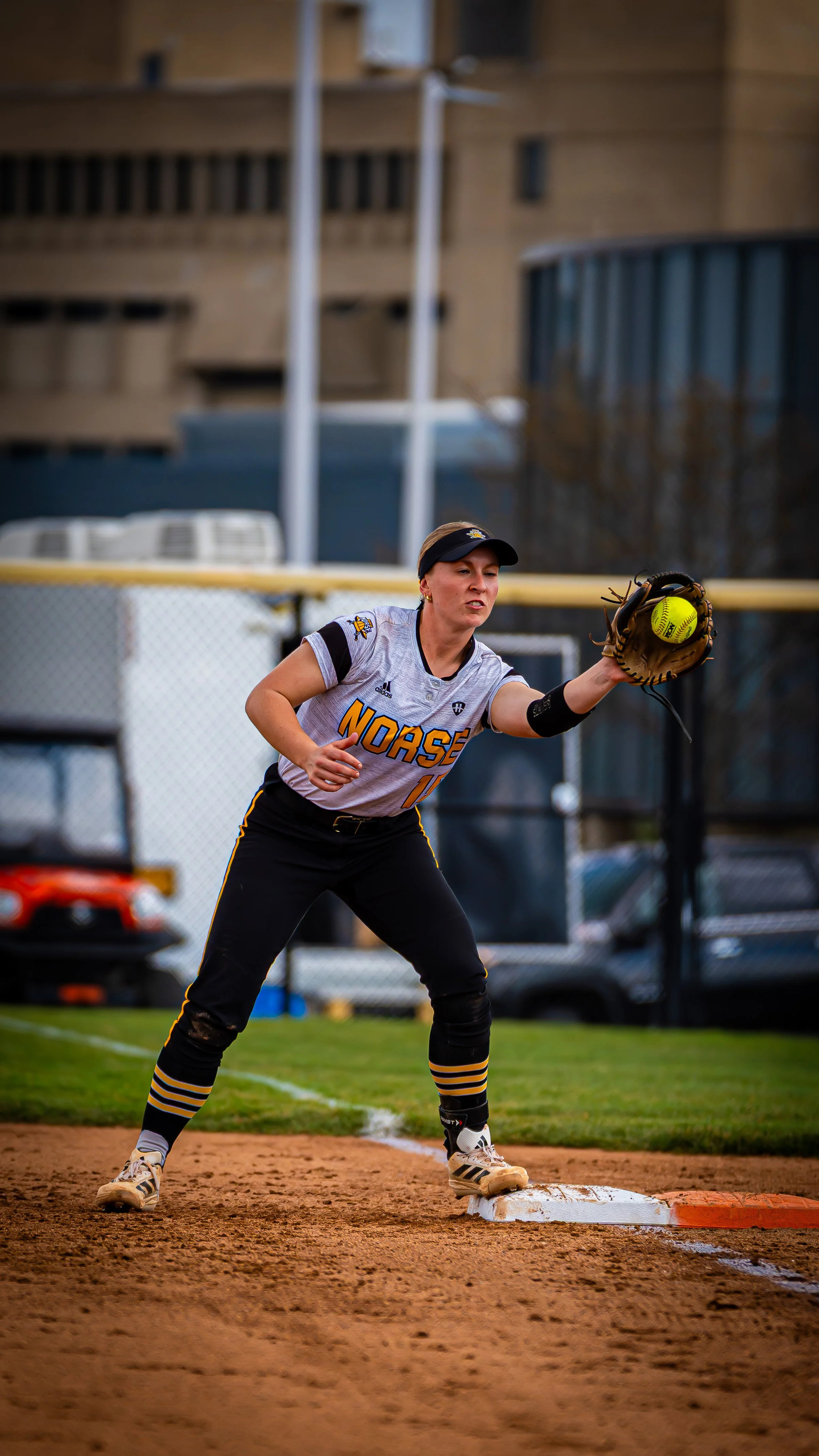 A female softball player wearing a gray jersey with 'Norse' written on it, black pants, striped knee-high socks, and a visor, catching a yellow softball in her glove at second base during a game.