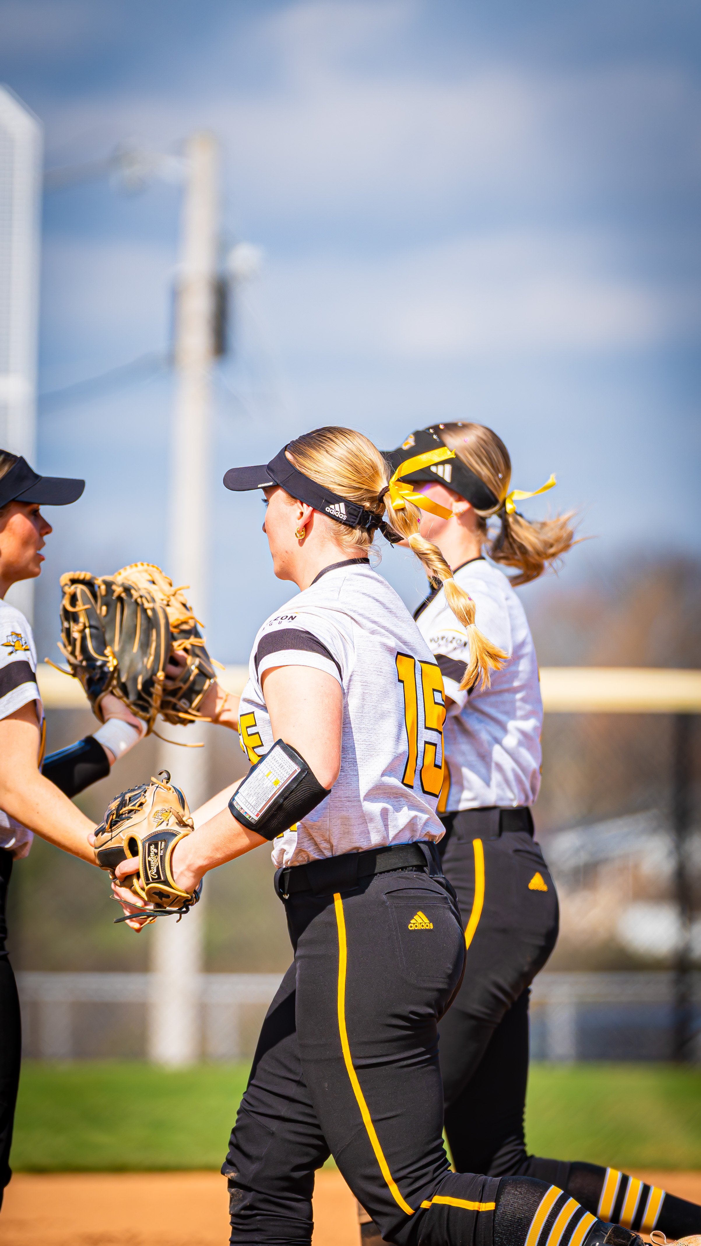 Softball players talking on the field wearing black and yellow uniforms, some with gloves and sunglasses.