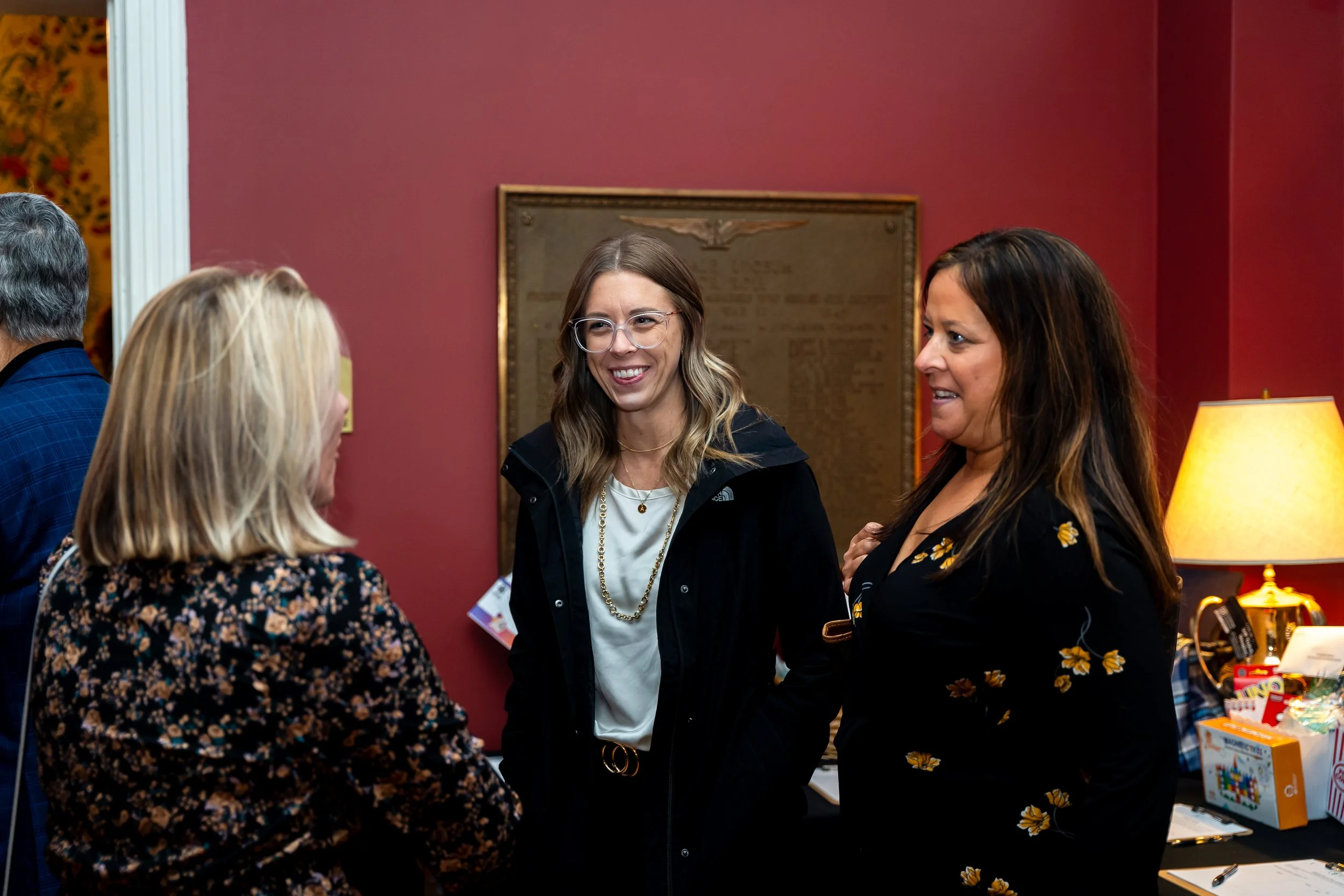 People engaged in conversation at an indoor event, with a woman wearing glasses and a black jacket smiling.