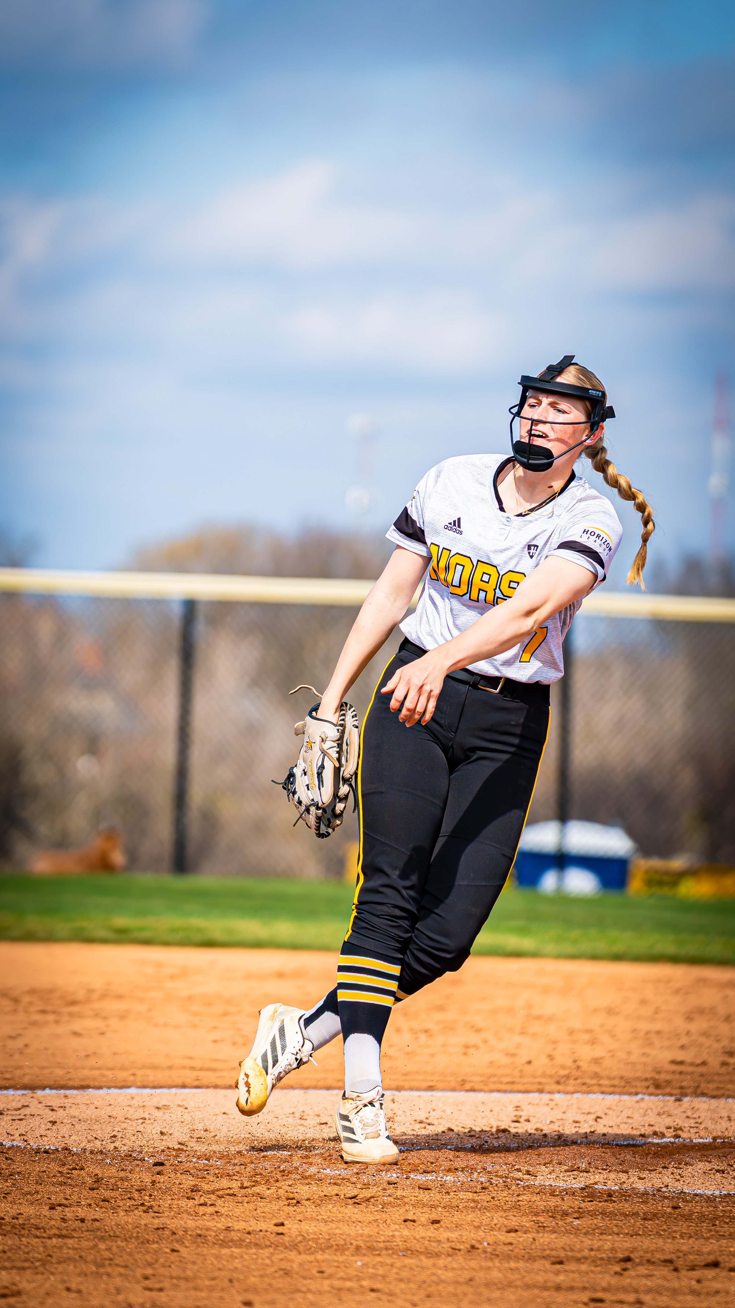 A female softball player on the field during a game, in a white and black uniform with yellow accents, wearing a face mask and holding a glove.
