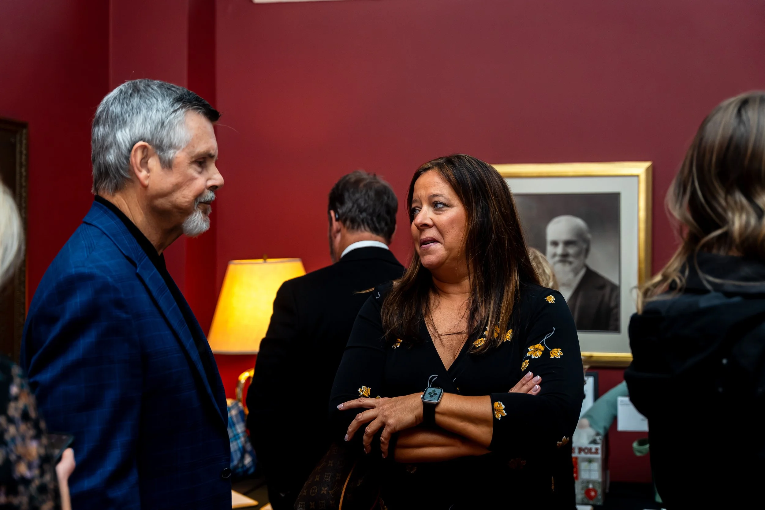Two women are engaged in conversation at a social gathering. The woman on the left has long dark hair and is wearing a black dress with yellow floral patterns, and a smartwatch on her wrist. The woman on the right has shoulder-length hair and is wear