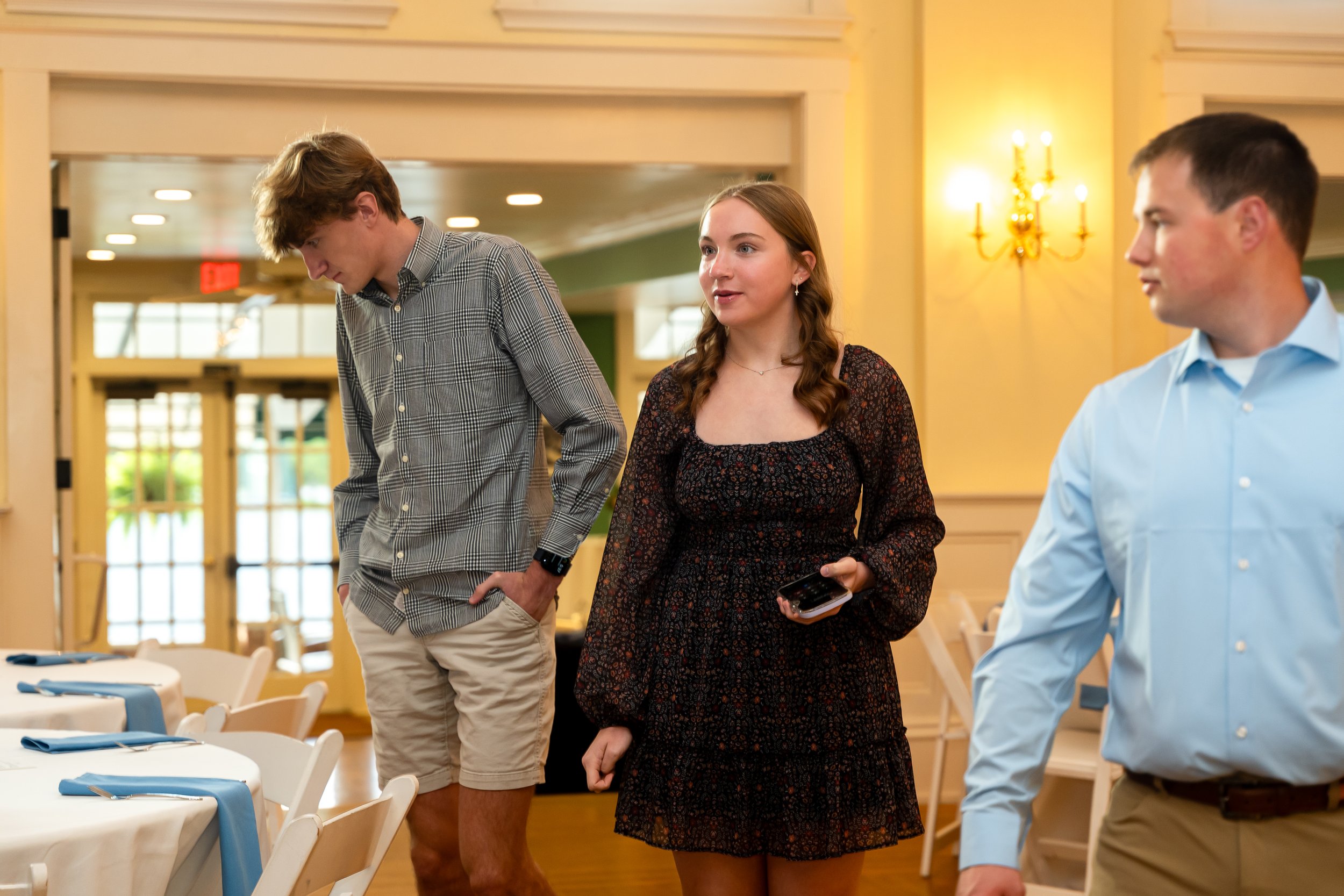 Three young adults at a social gathering, standing near a table with blue napkins, in a well-lit room with yellow walls and a chandelier. The woman in the center is holding a phone, the man on the left is looking down, and the man on the right is loo