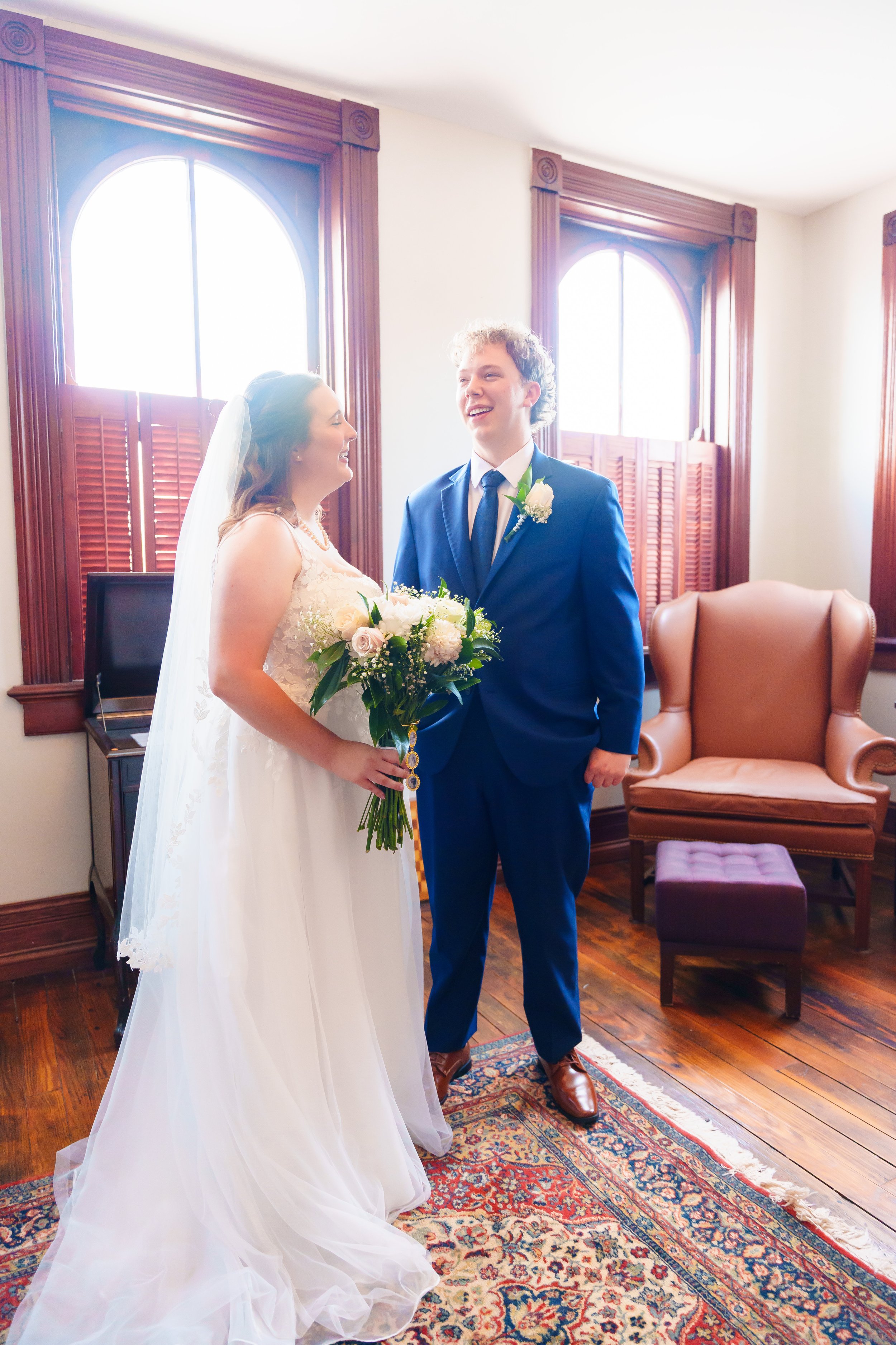 A bride and groom standing together inside a room with wooden window frames, smiling and looking at each other. The bride wears a white wedding dress and veil, holding a bouquet of white and pink flowers. The groom wears a blue suit with a white shir