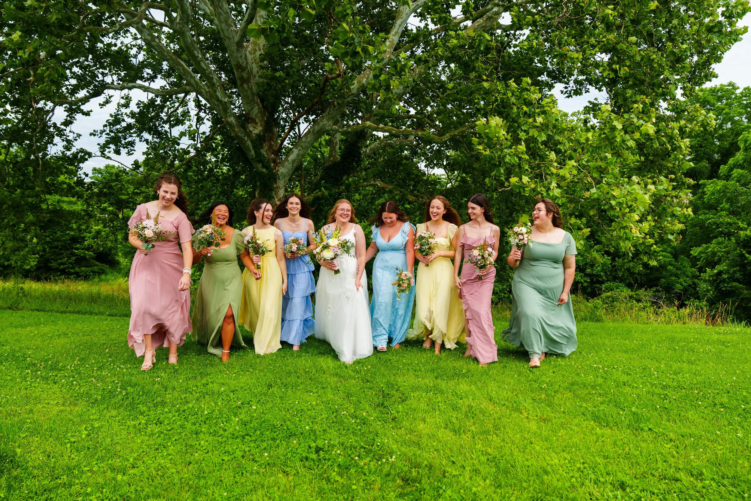 Group of women in colorful dresses walking and talking outdoors under a large tree, smiling and holding flowers.