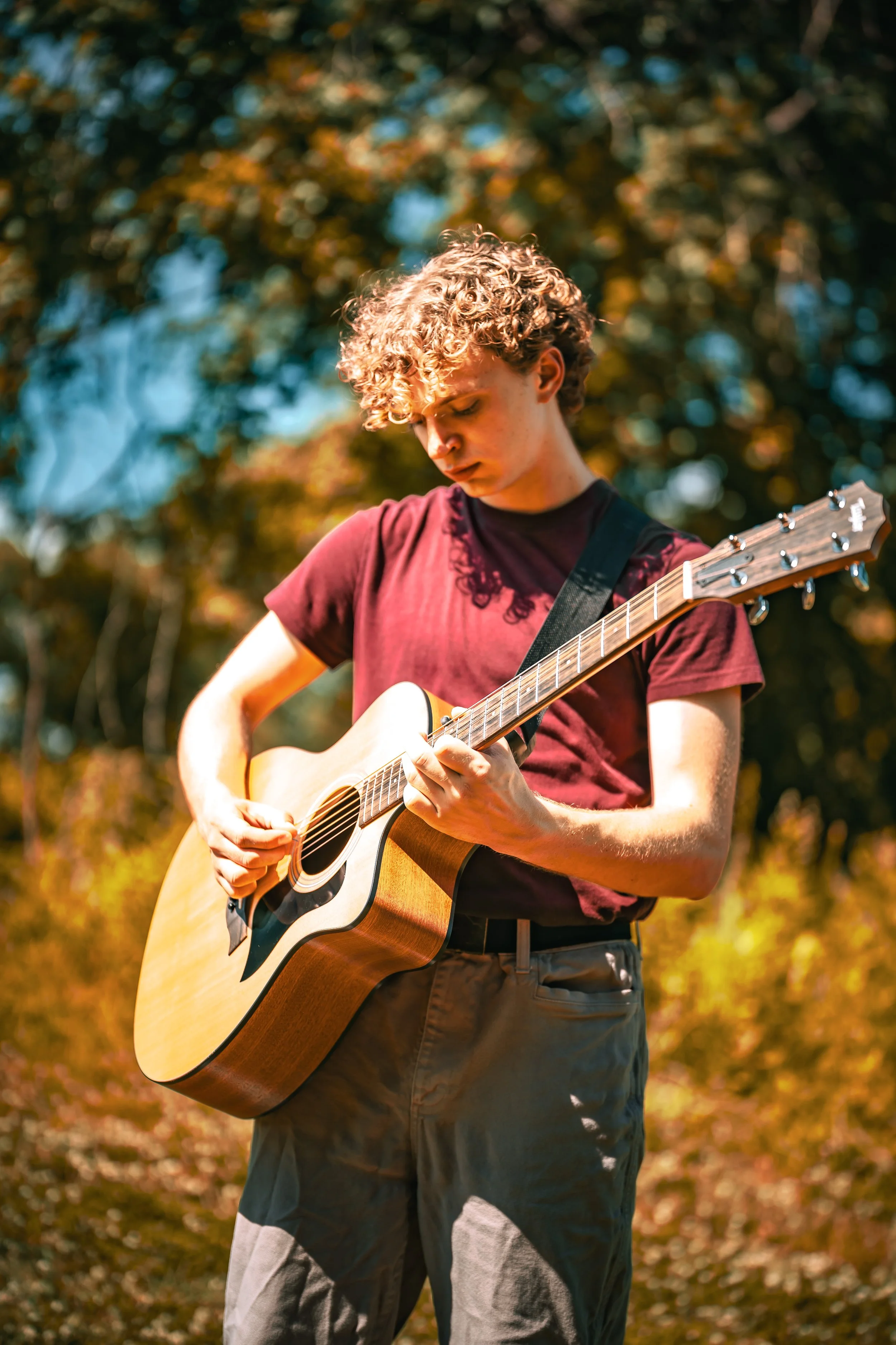Young man with curly hair playing an acoustic guitar outdoors during autumn.