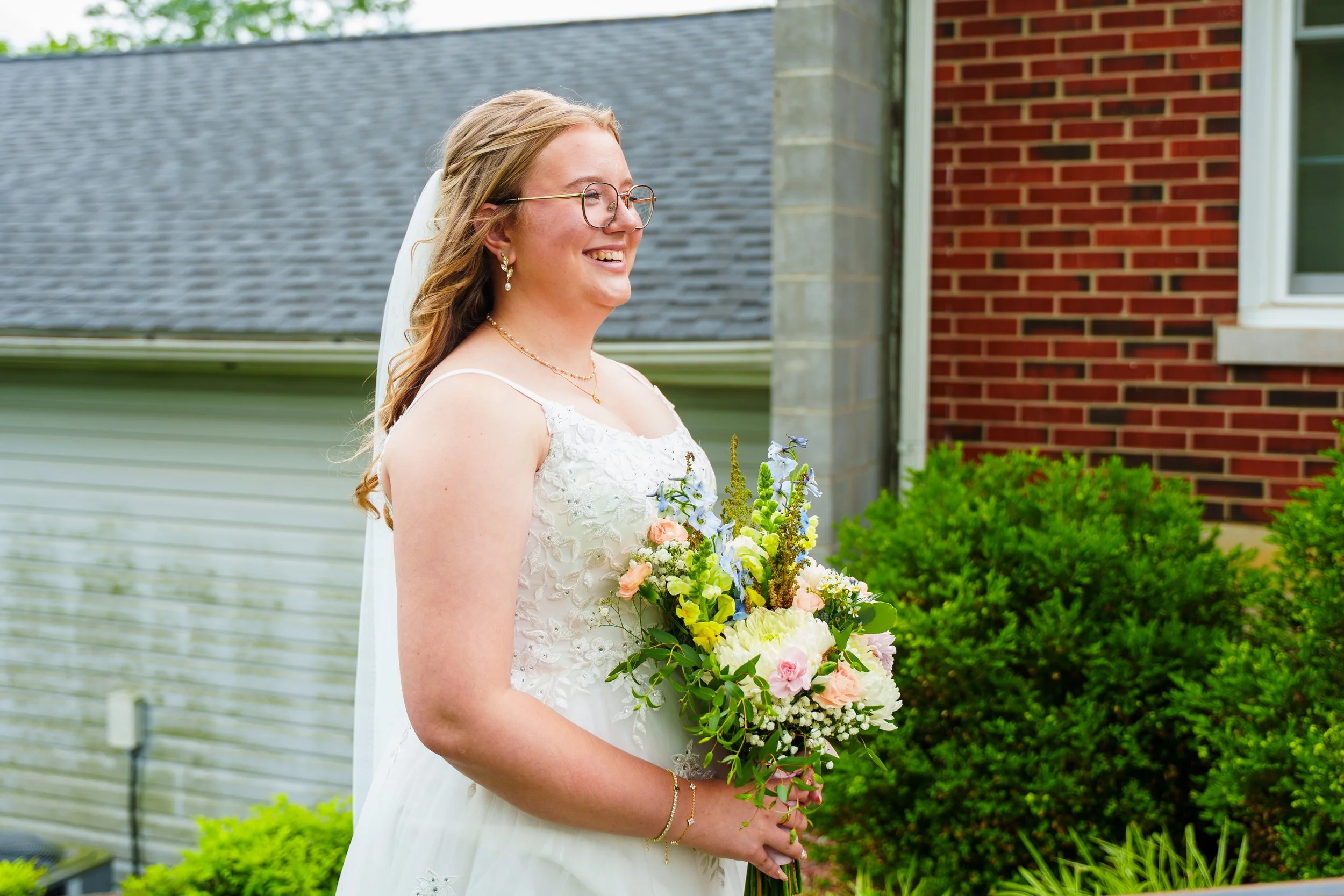 A bride with glasses and earrings smiling and holding a bouquet of flowers outdoors in front of a building with a brick wall and greenery.