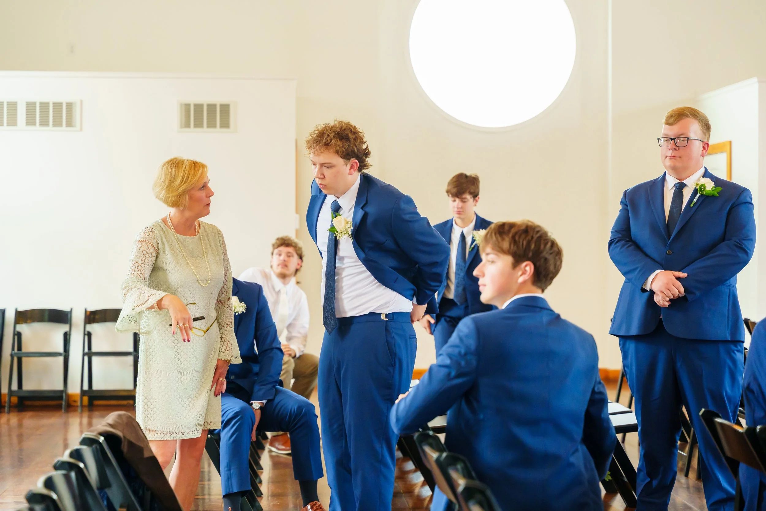 A woman in a white lace dress and pearl necklace is speaking to a young man in a blue suit at a formal event. Several other young men in blue suits are seated or standing nearby in a spacious room with large circular window and chairs arranged in row