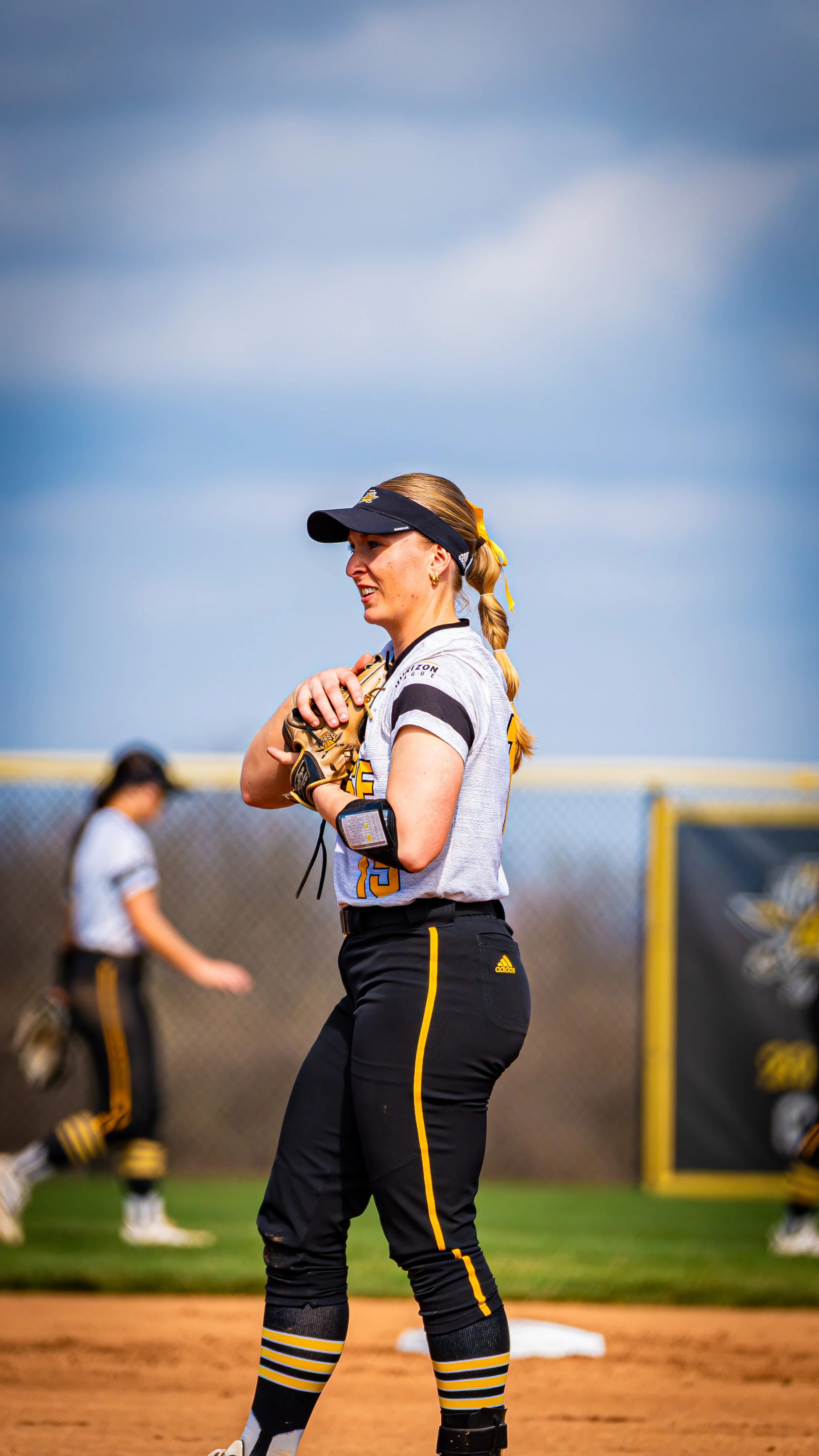 A female softball player standing on the field, wearing a white jersey with black and yellow details, black pants with yellow stripes, and a black cap, holding a softball glove, with other players and a chain-link fence in the background.