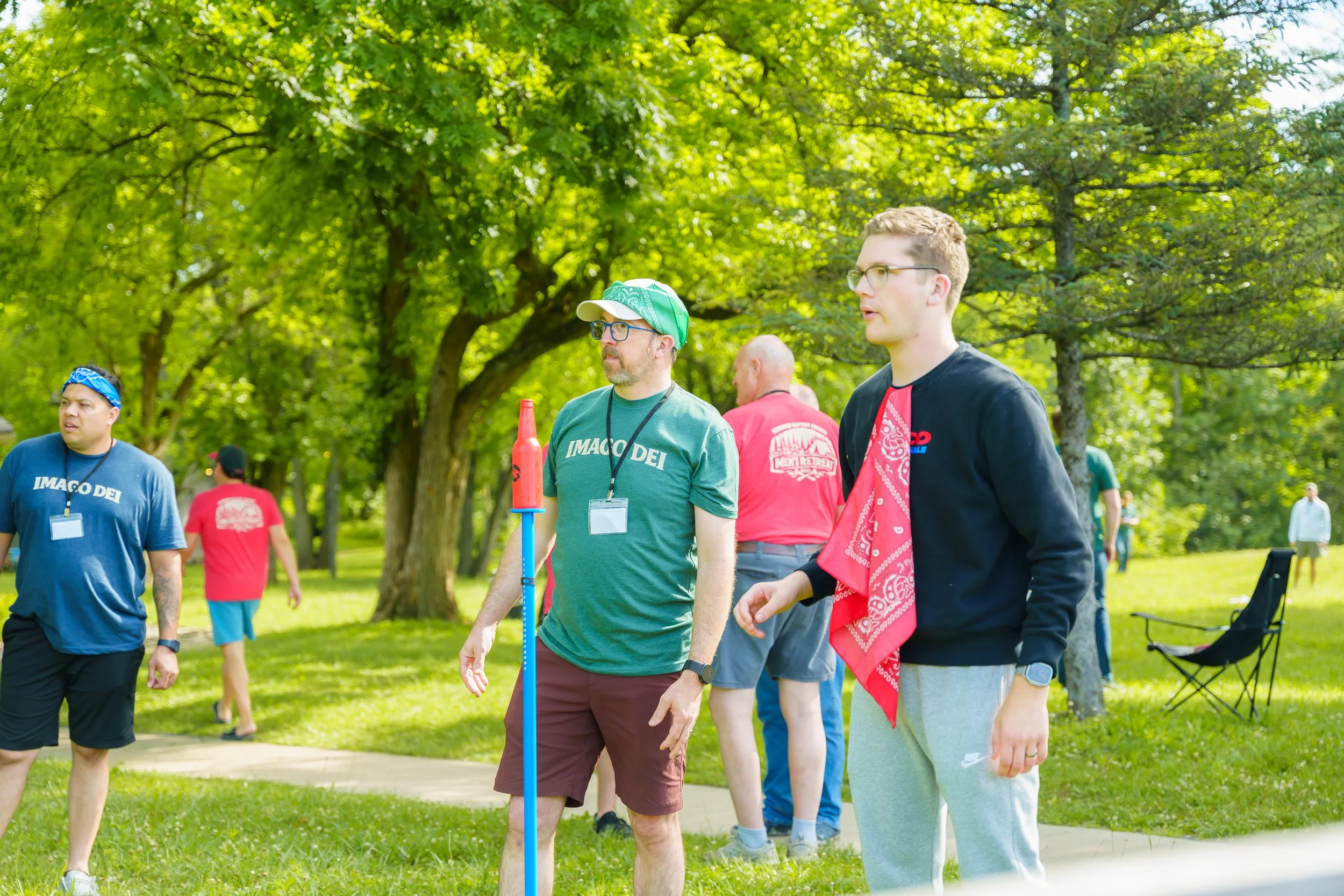 Group of people outdoors in a park with green trees, some appear to be participating in an event, wearing casual clothing and name tags.