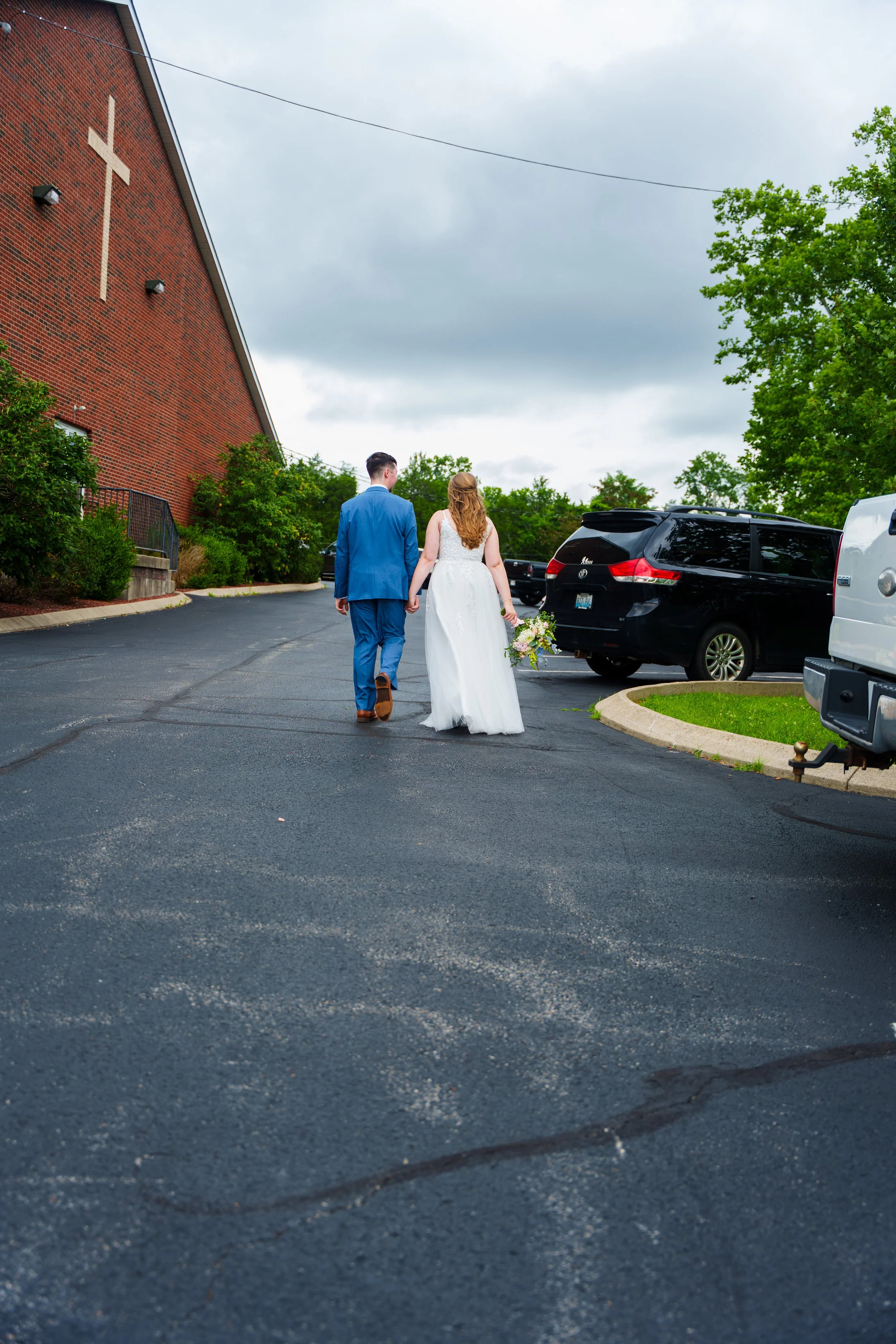 A couple dressed in wedding attire walking hand in hand in a parking lot, with cars and a brick building with a cross on it in the background, under a cloudy sky.