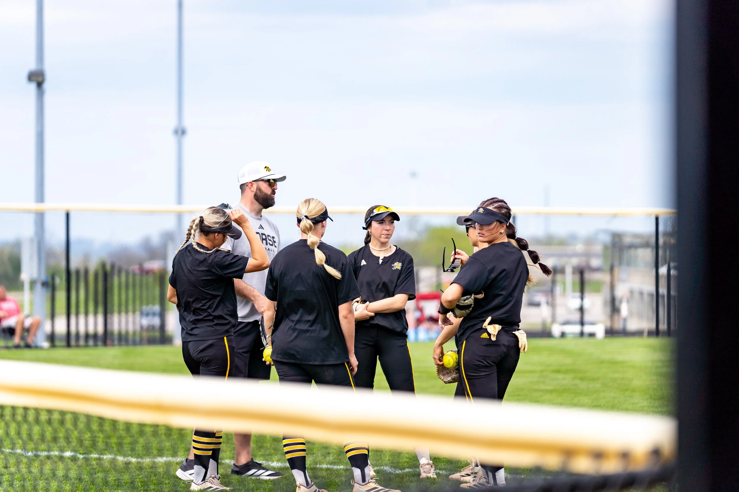 A group of women and a man in black and yellow sportswear standing on a sports field during daytime, likely women softball players with a coach or team leader.