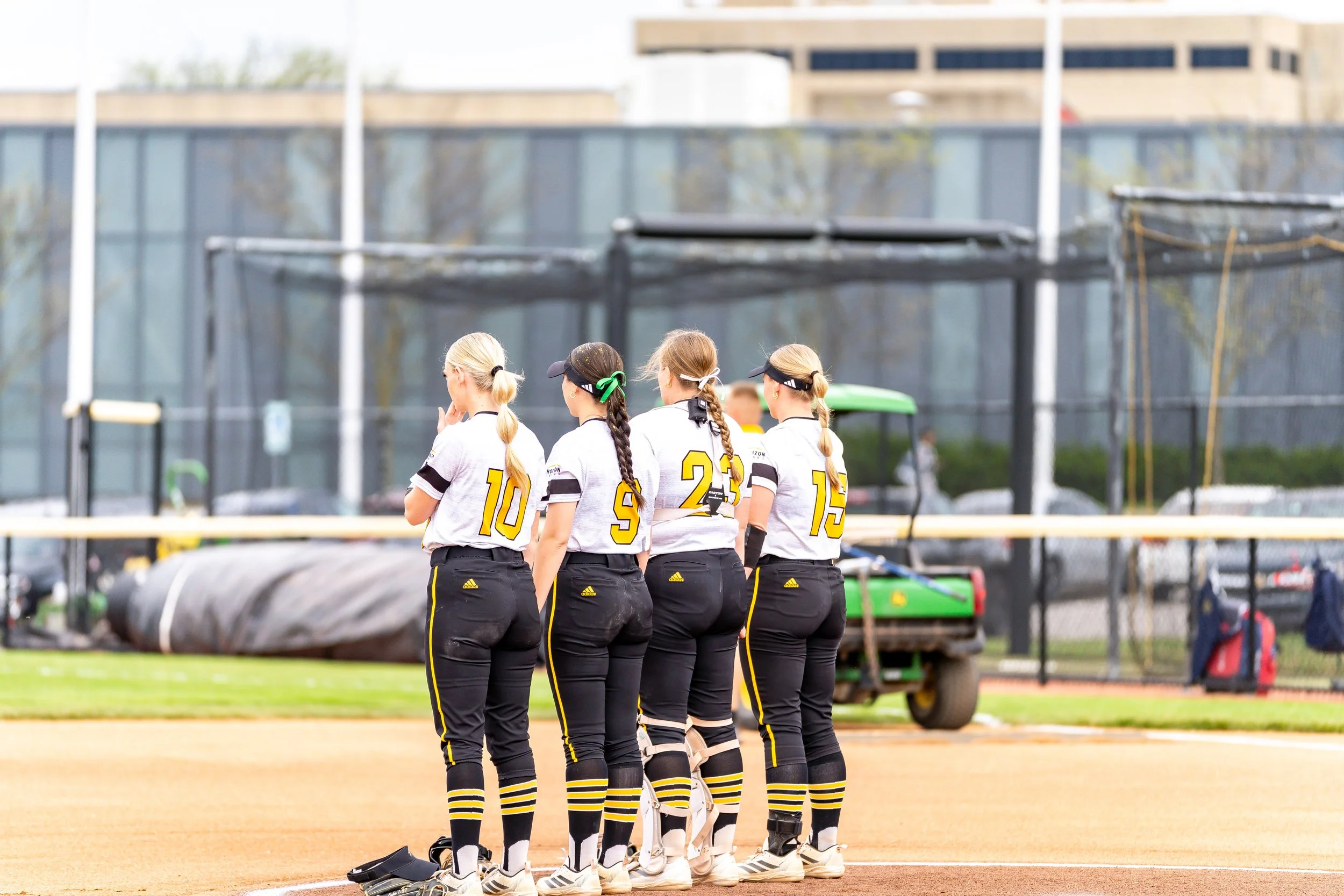 A group of female softball players standing on a softball field, wearing black and yellow uniforms, with their backs to the camera, with a building and sports equipment in the background.