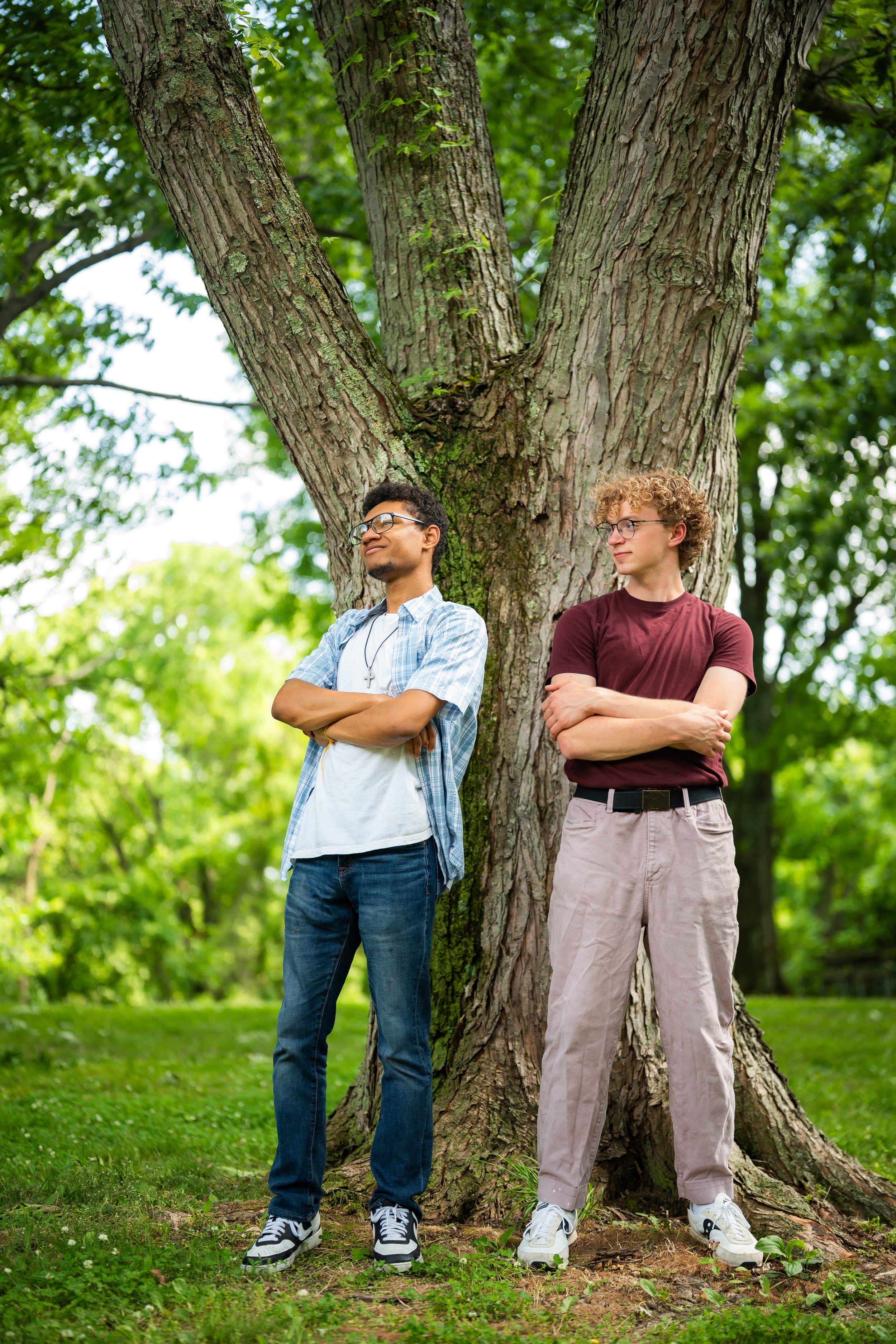 Two young men standing with arms crossed in front of a large tree in a park, surrounded by green foliage.