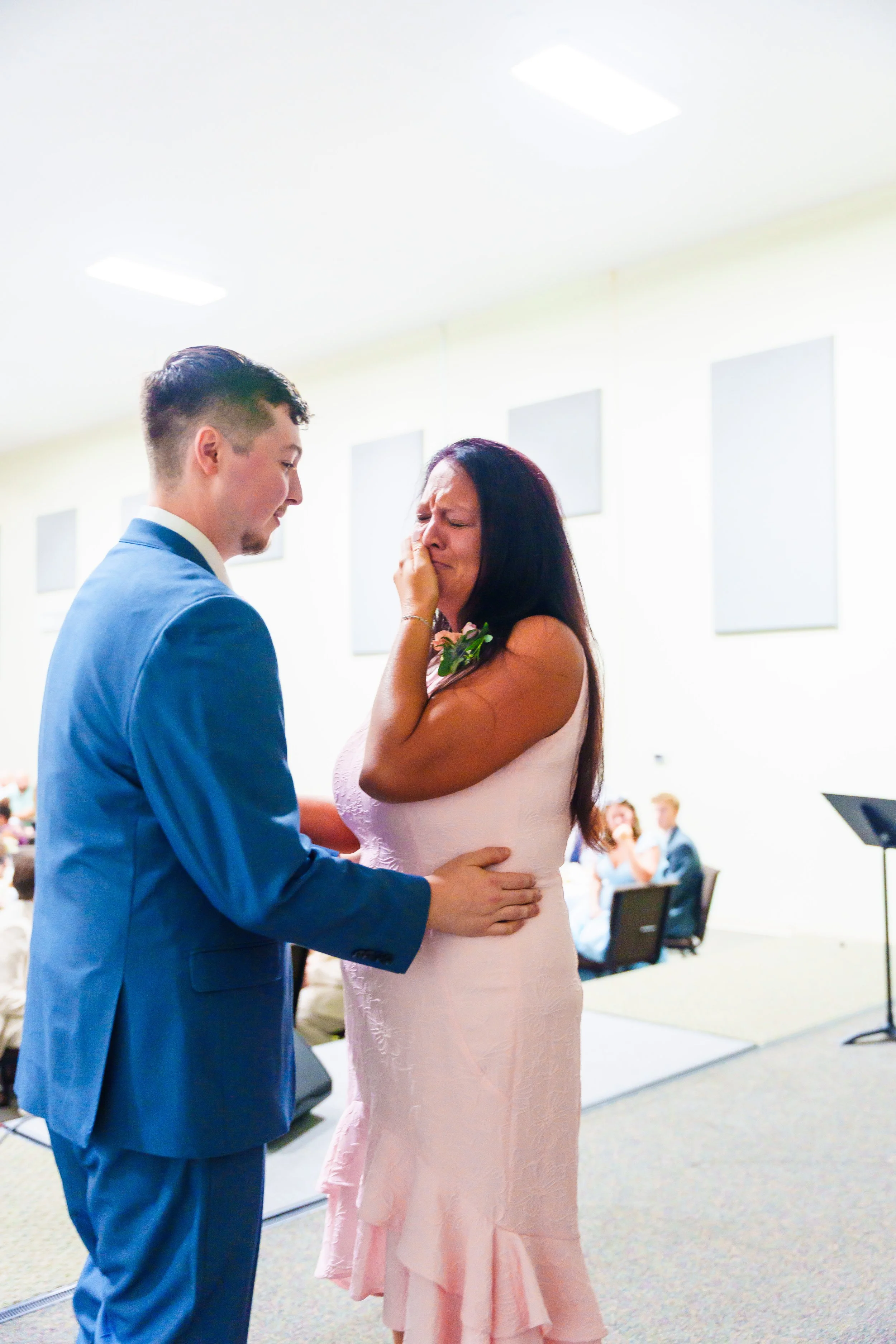 A couple dressed in formal attire, with the woman in a pink dress and the man in a blue suit, holding hands and embracing during a wedding ceremony in a brightly lit room, with seated guests in the background.