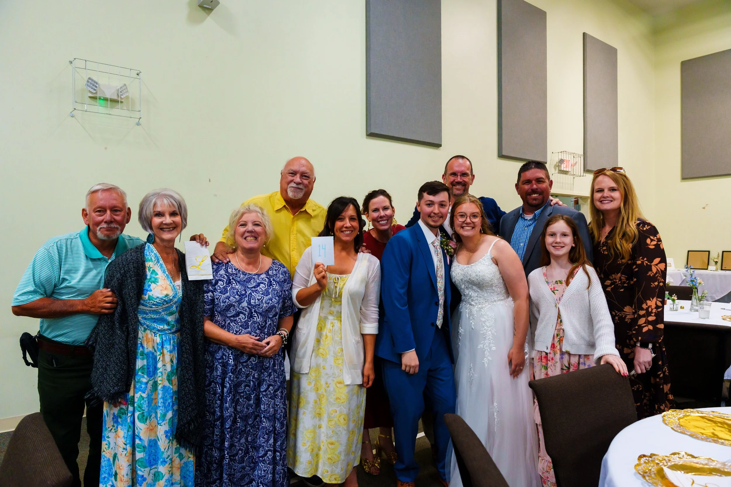 A group of diverse smiling people at a celebration or wedding, standing together indoors against a light green wall with soundproof panels. Some are holding small signs, and a table with yellow and white decorations is in the foreground.