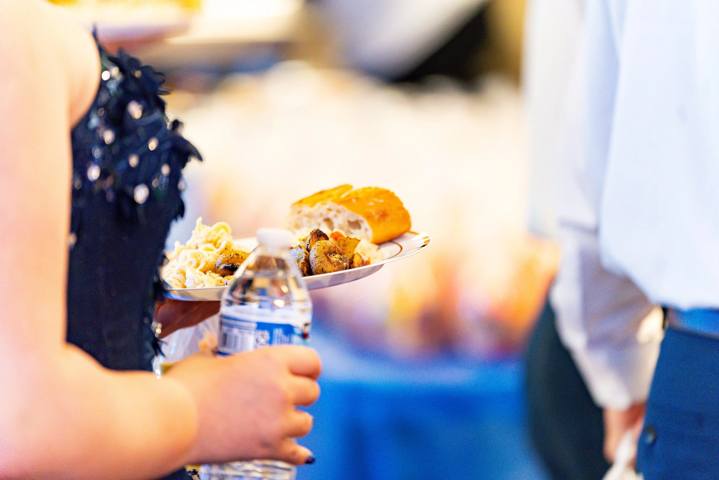 Person holding a white plate with assorted food, including bread and mushrooms, near a water bottle, with other people in the background.
