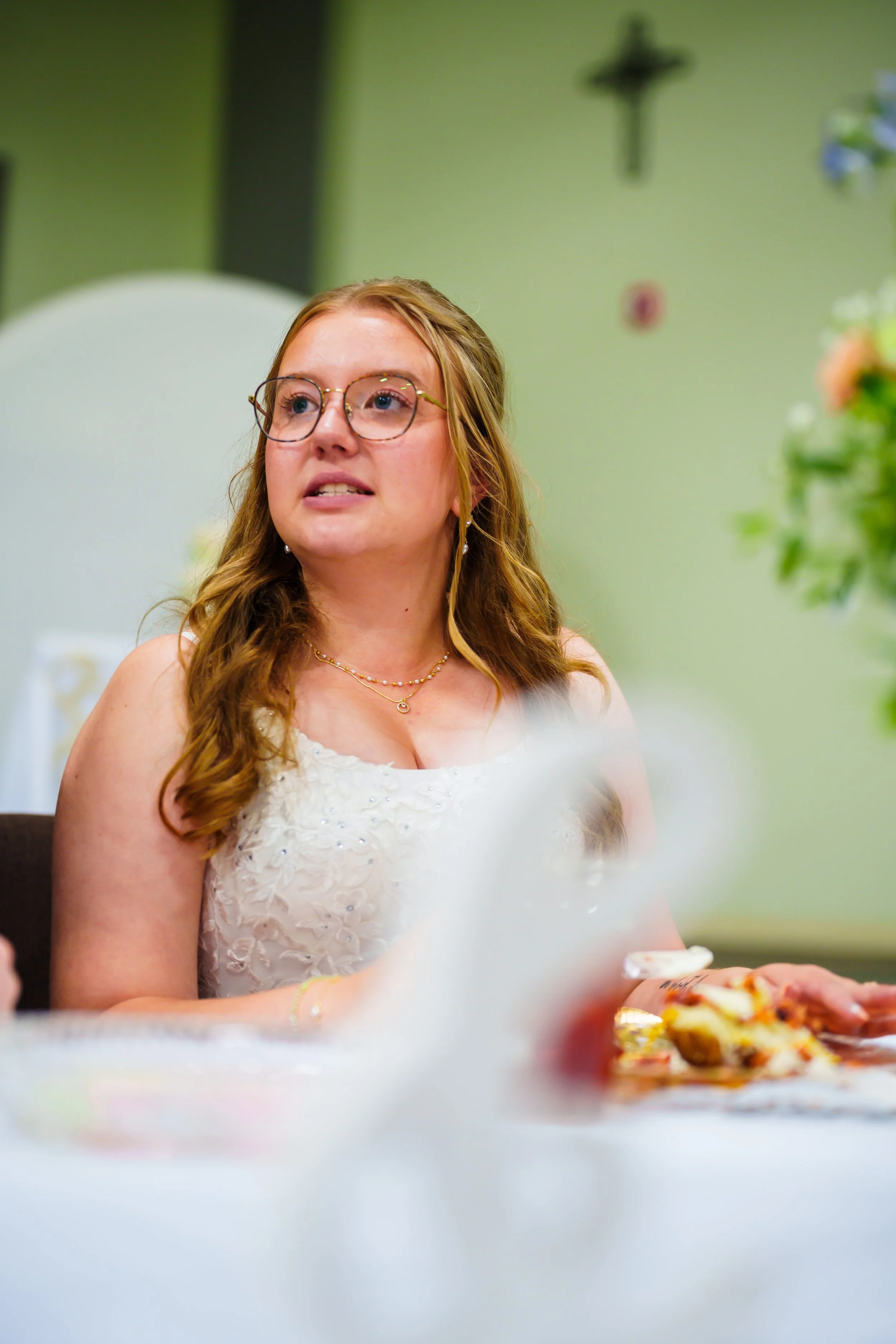 A woman with long wavy blonde hair and glasses wearing a white dress, sitting at a table with a plate of food, in a room with green walls and floral decorations.