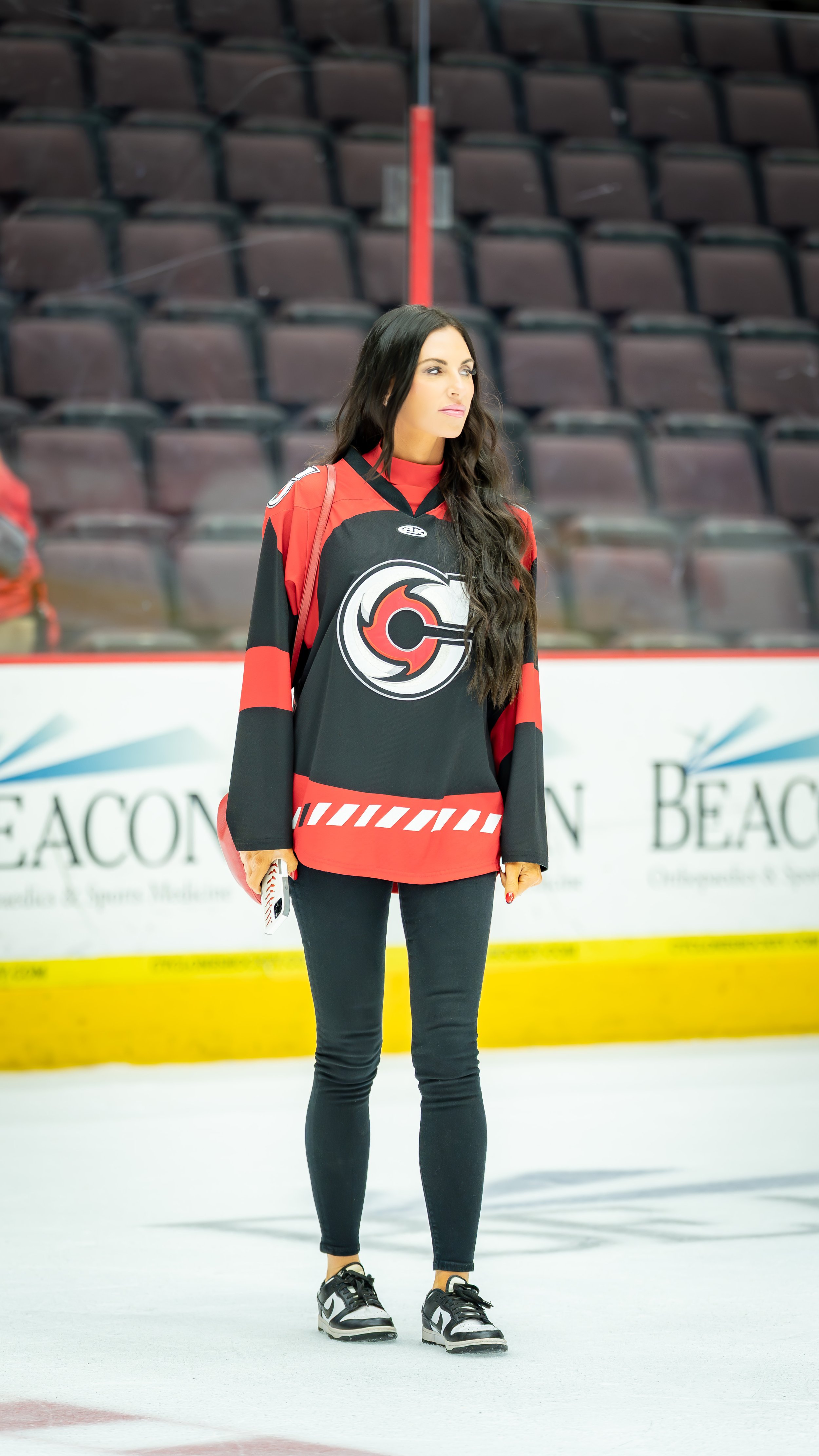 A woman standing on an ice rink wearing a black and red hockey jersey with a large logo on the front, black pants, and black and white sneakers. She has long dark hair and appears to be focused, with empty stadium seats in the background.