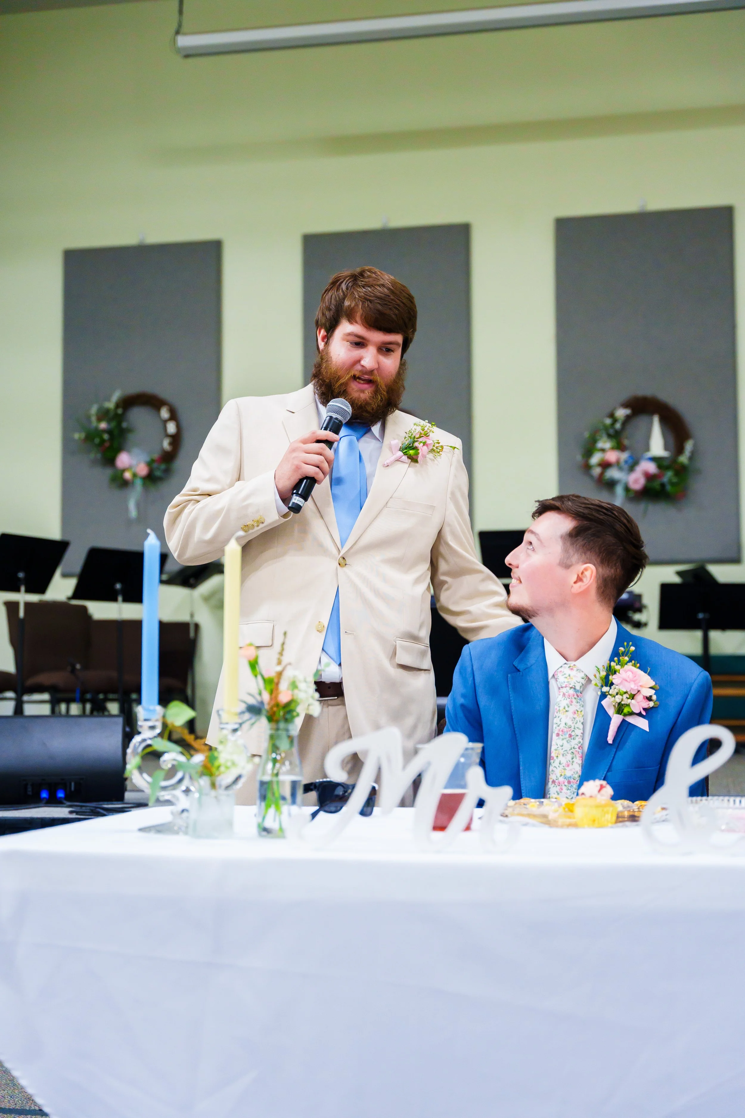 A man with a beard in a beige suit is holding a microphone and speaking to another man in a blue suit seated at a decorated table during a celebration or wedding event at an indoor venue.