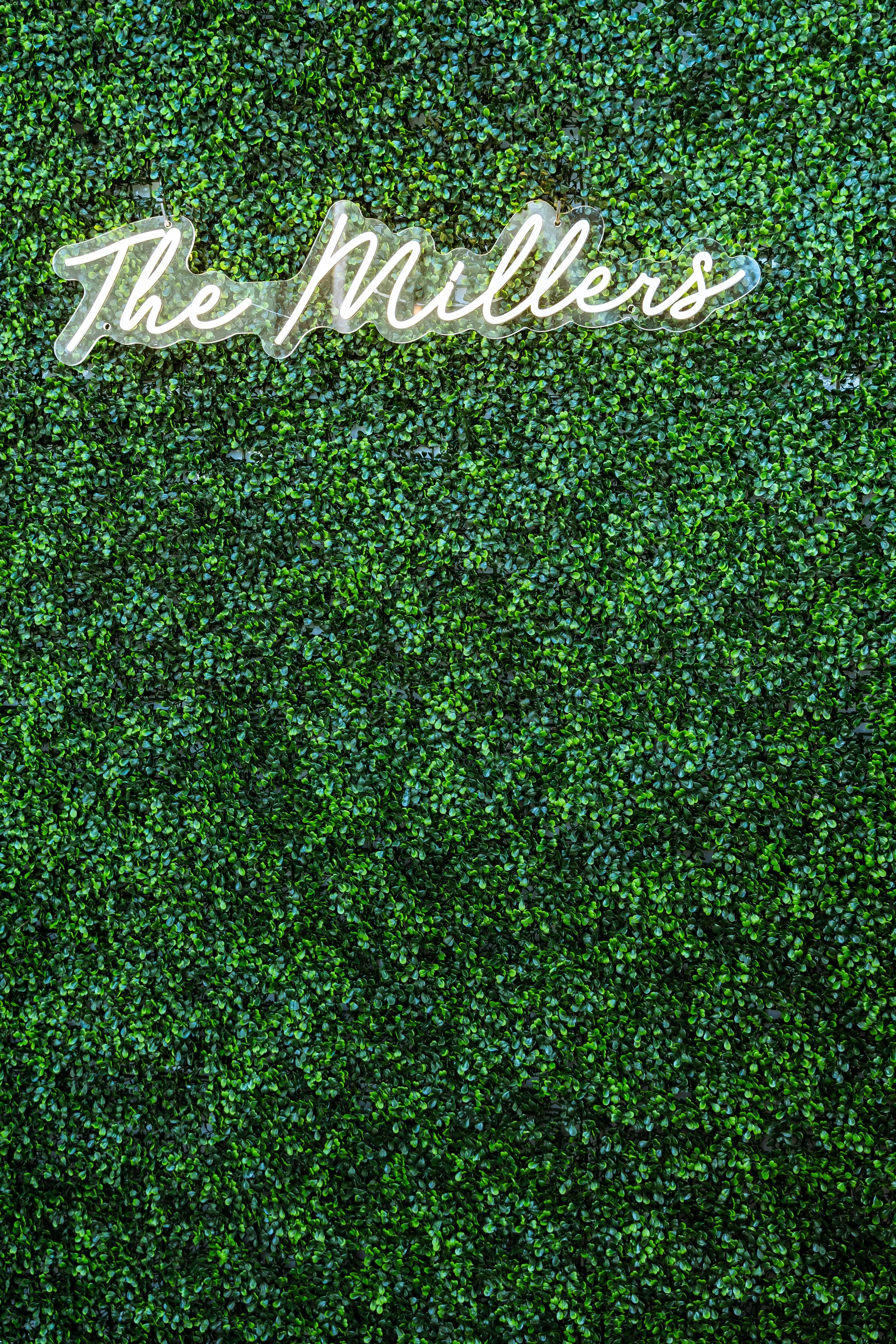 Neon sign that says 'The Mills' mounted on a lush green artificial foliage wall.