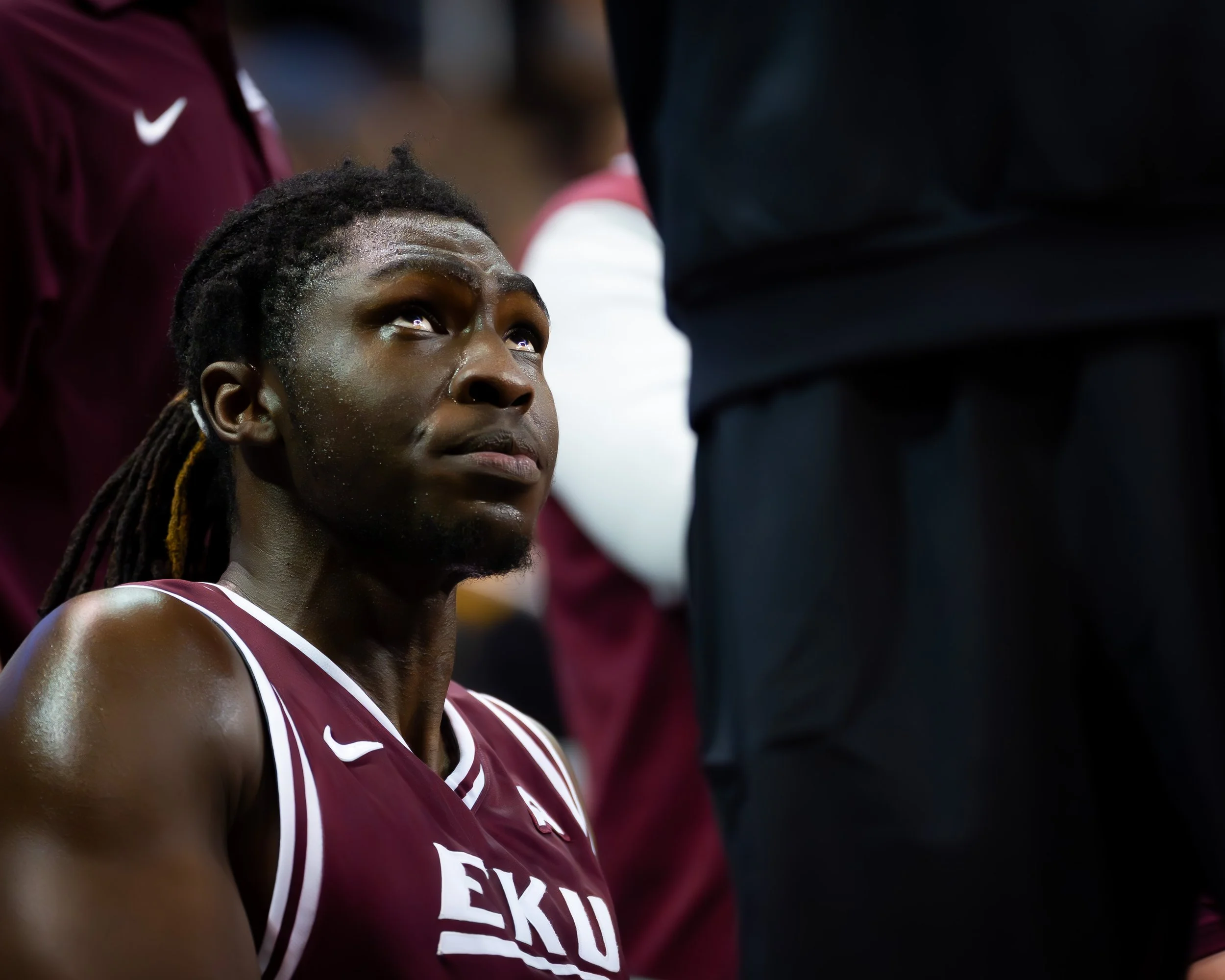 A male basketball player with dreadlocks and sweat on his face, wearing a maroon jersey with white lettering, sitting on the sideline and looking up.